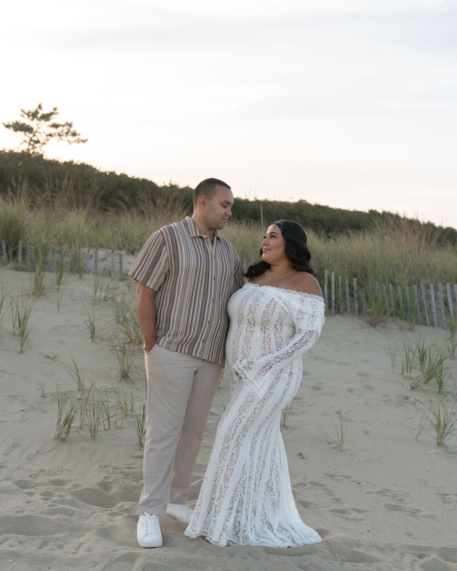 Couple poses on sandy beach dunes at sunset, woman in white lace off-shoulder gown, man in striped shirt and khakis.