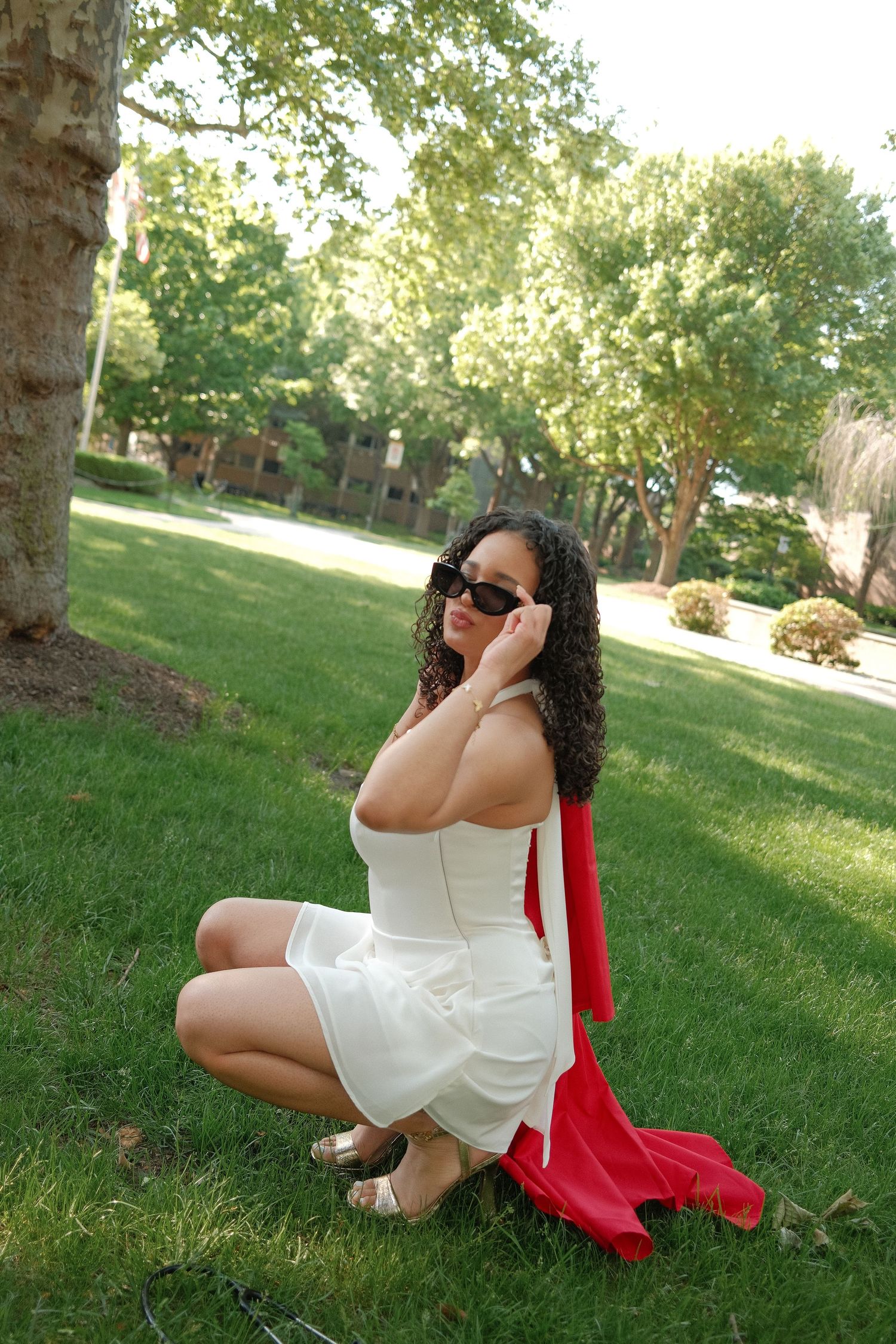 Young woman in white dress and sunglasses poses on grass with red graduation gown.