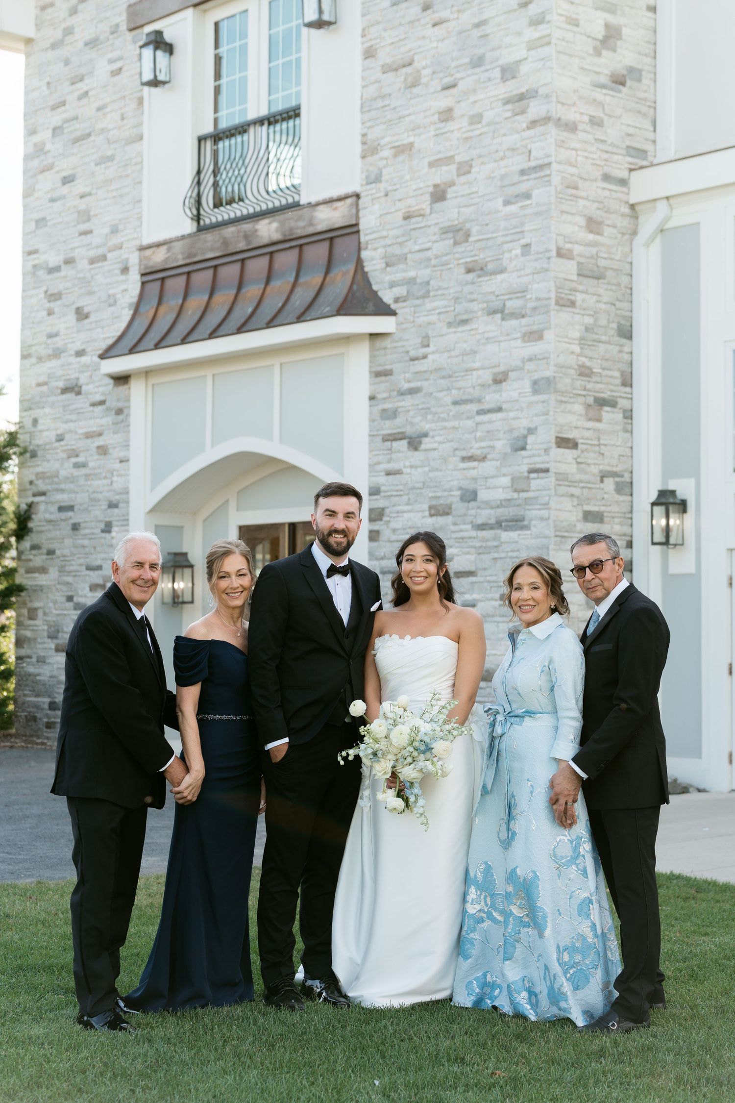 Wedding party family portrait outside stone venue with bride, groom, and parents in formal attire.