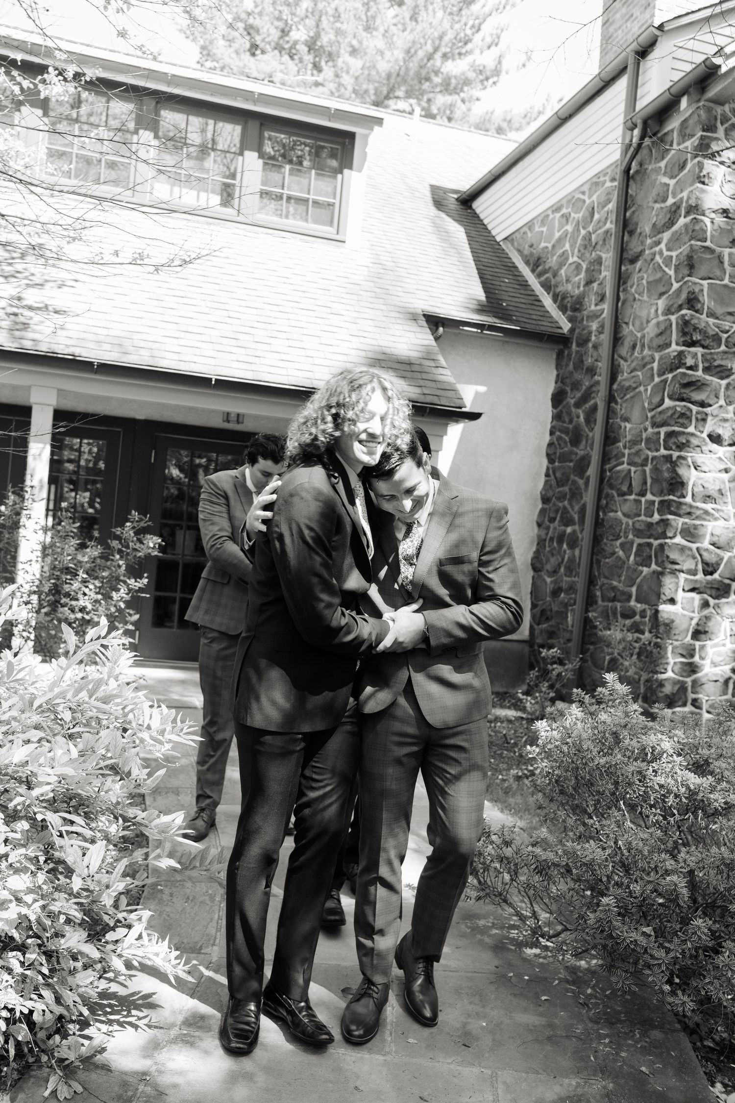 Groomsmen in dark suits playfully posing together outside a stone building in black and white.