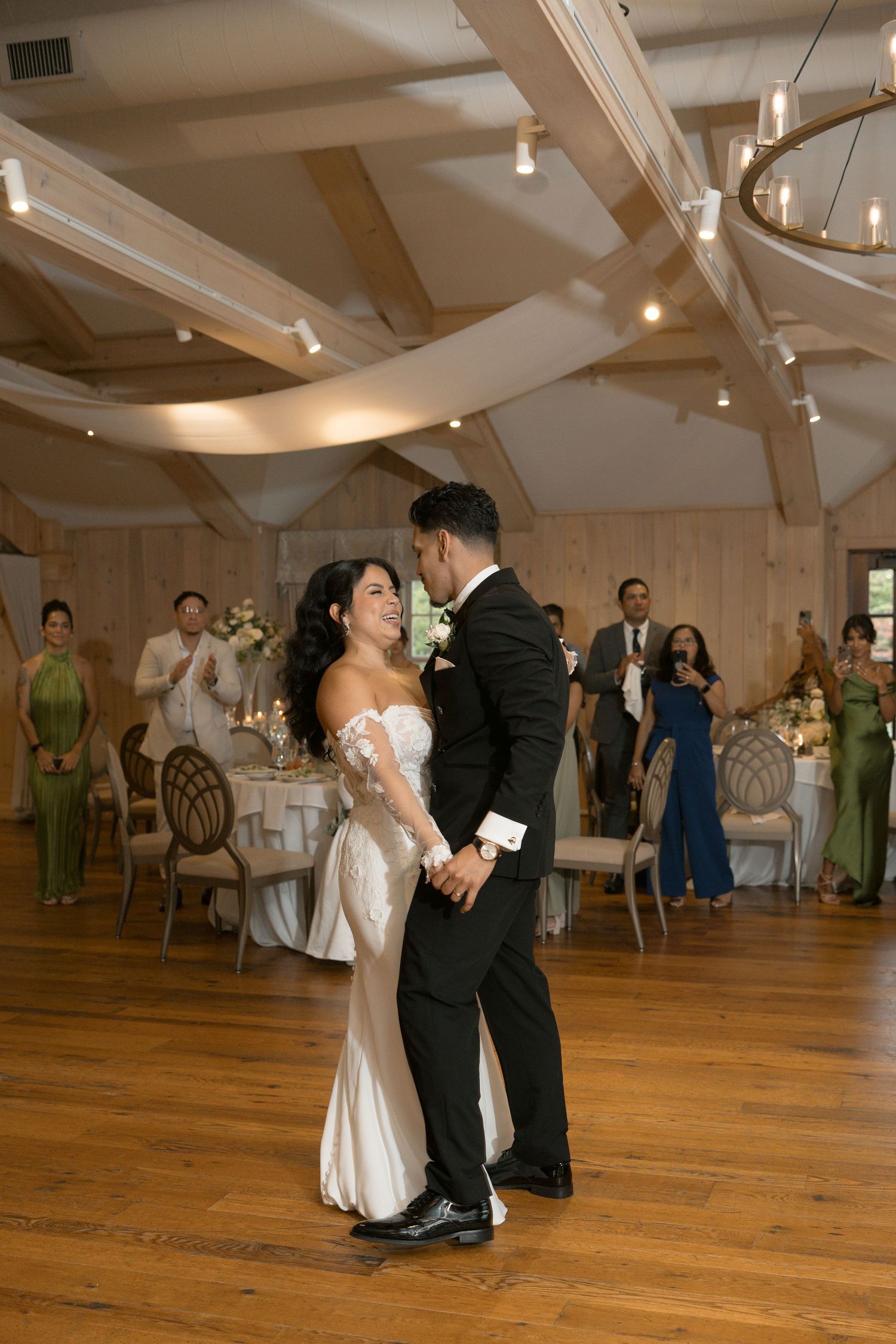Bride and groom share their first dance in an elegant wood-beamed reception hall while guests applaud around them.