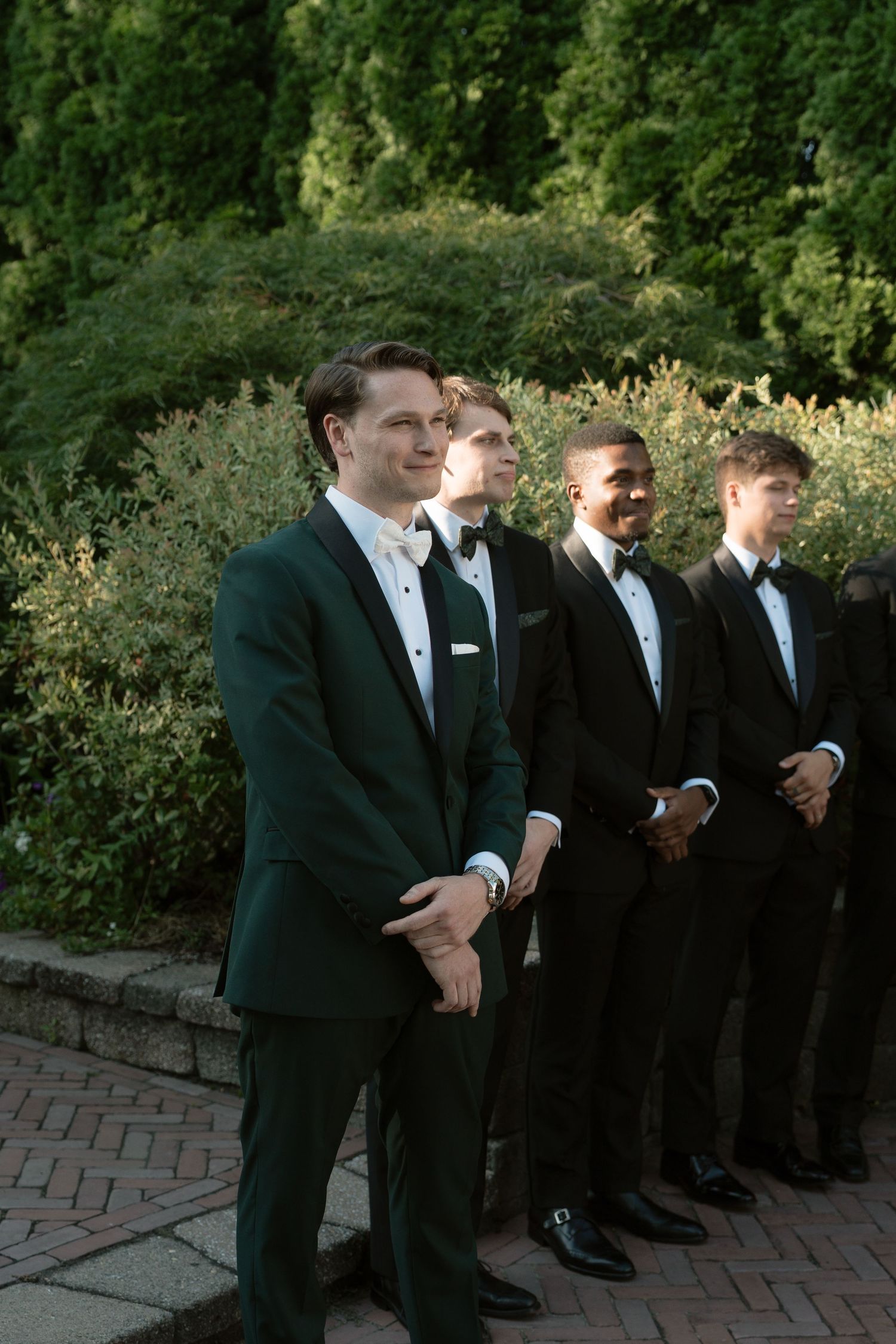 Groom in dark green tuxedo stands with groomsmen in black suits in a lush garden setting.