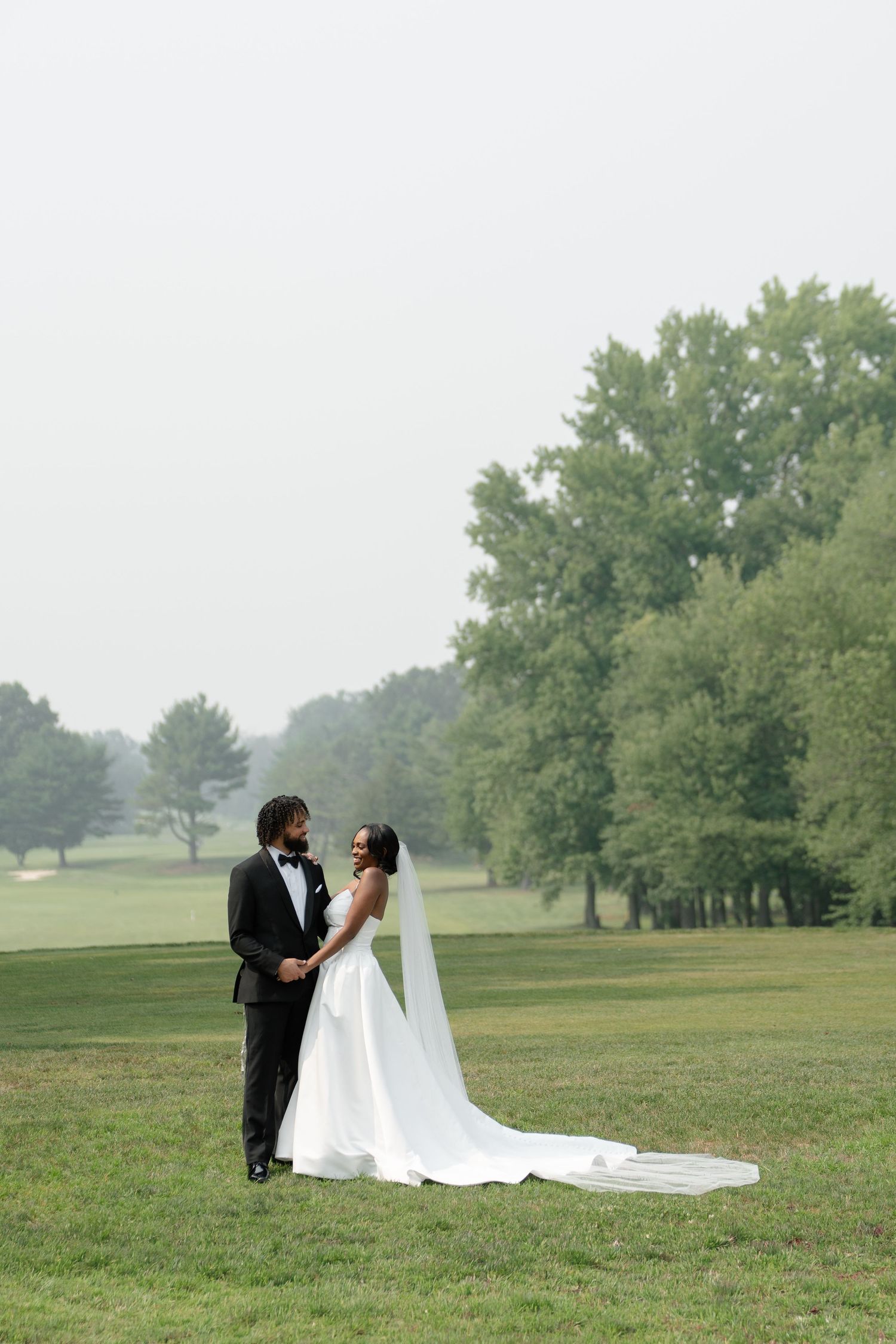 A bride and groom share a romantic moment on a lush green lawn, with the bride's long veil flowing behind her.