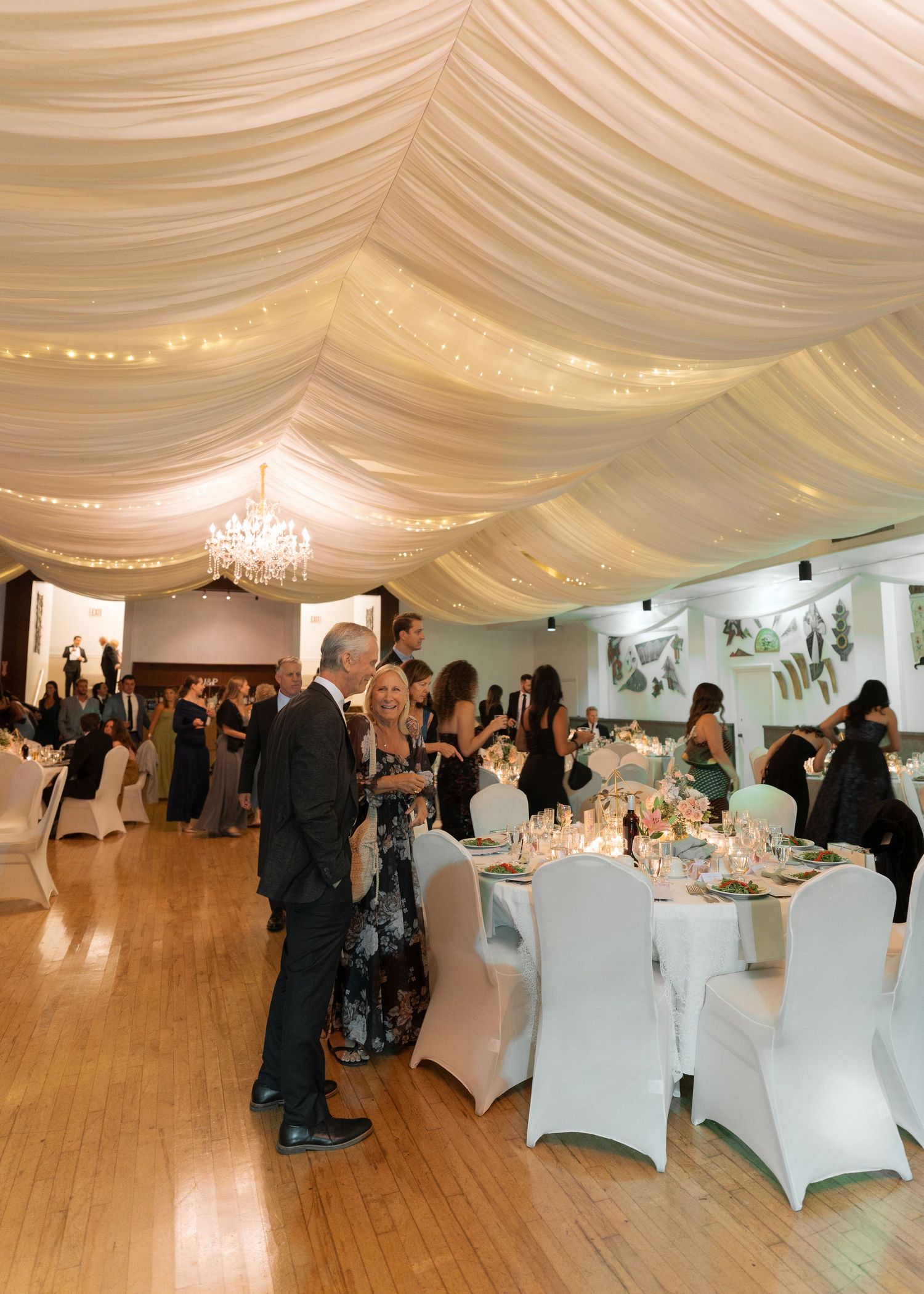 Beautiful wedding reception venue with draped white ceiling, chandeliers, round tables, and dancing guests.