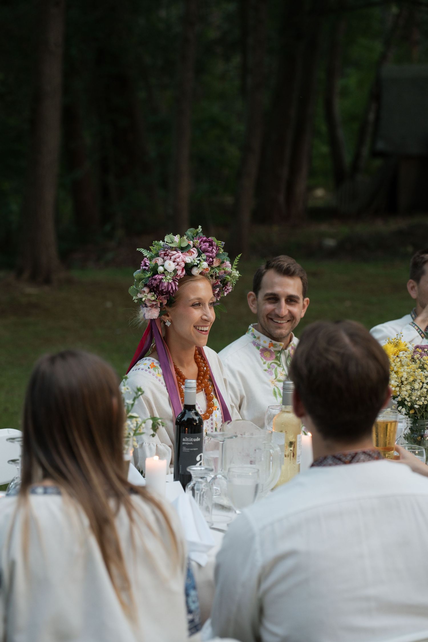 Woman wearing floral crown at outdoor garden dinner party with guests seated at decorated table with candles.