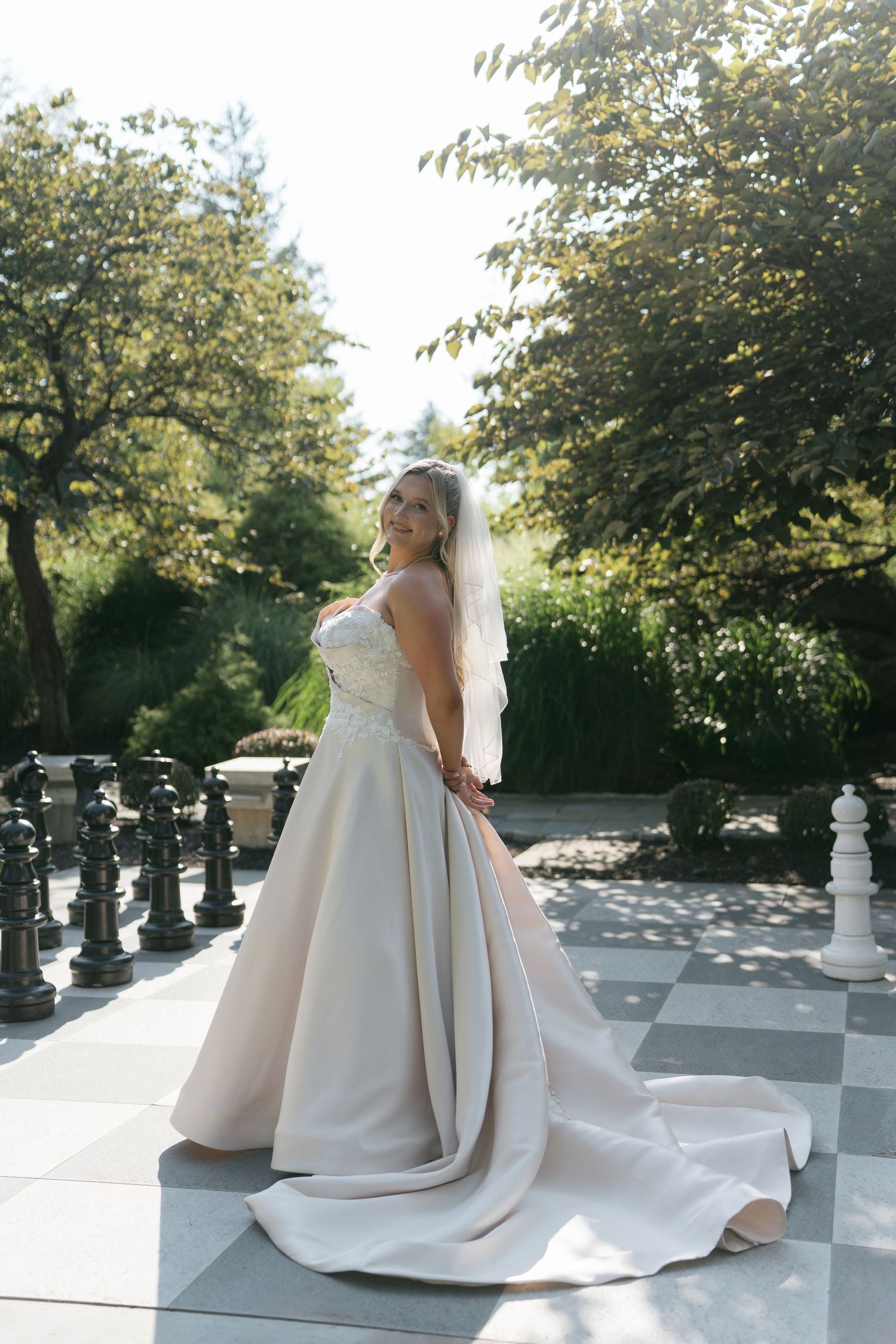 Bride in a strapless ball gown with lace bodice and long train poses on a checkered patio surrounded by lush greenery.