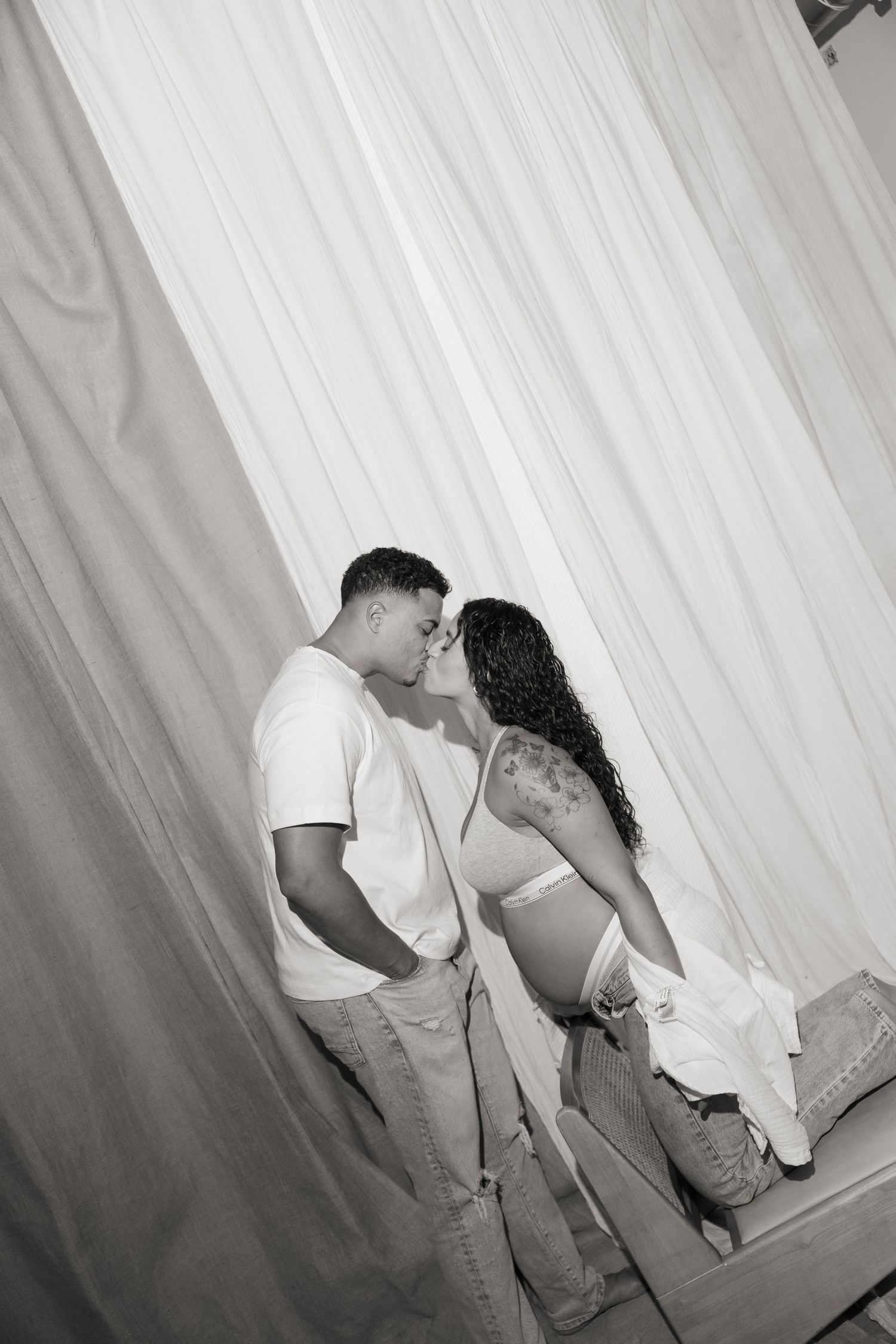 Black and white photo of a couple sharing a tender kiss in front of sheer white curtains, woman showing baby bump.