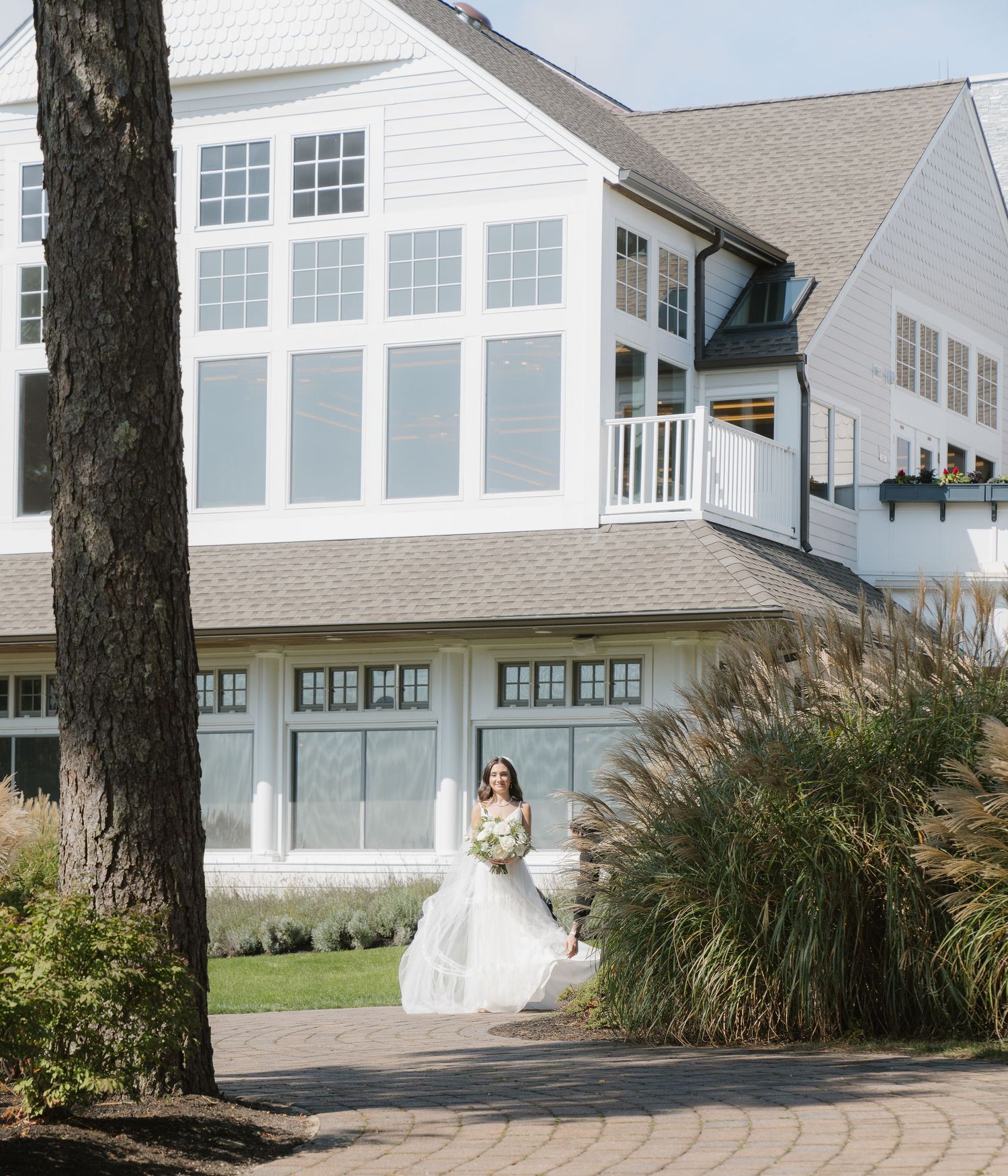 A bride in a white wedding dress poses in front of a coastal beach house with ornamental grasses in the foreground.