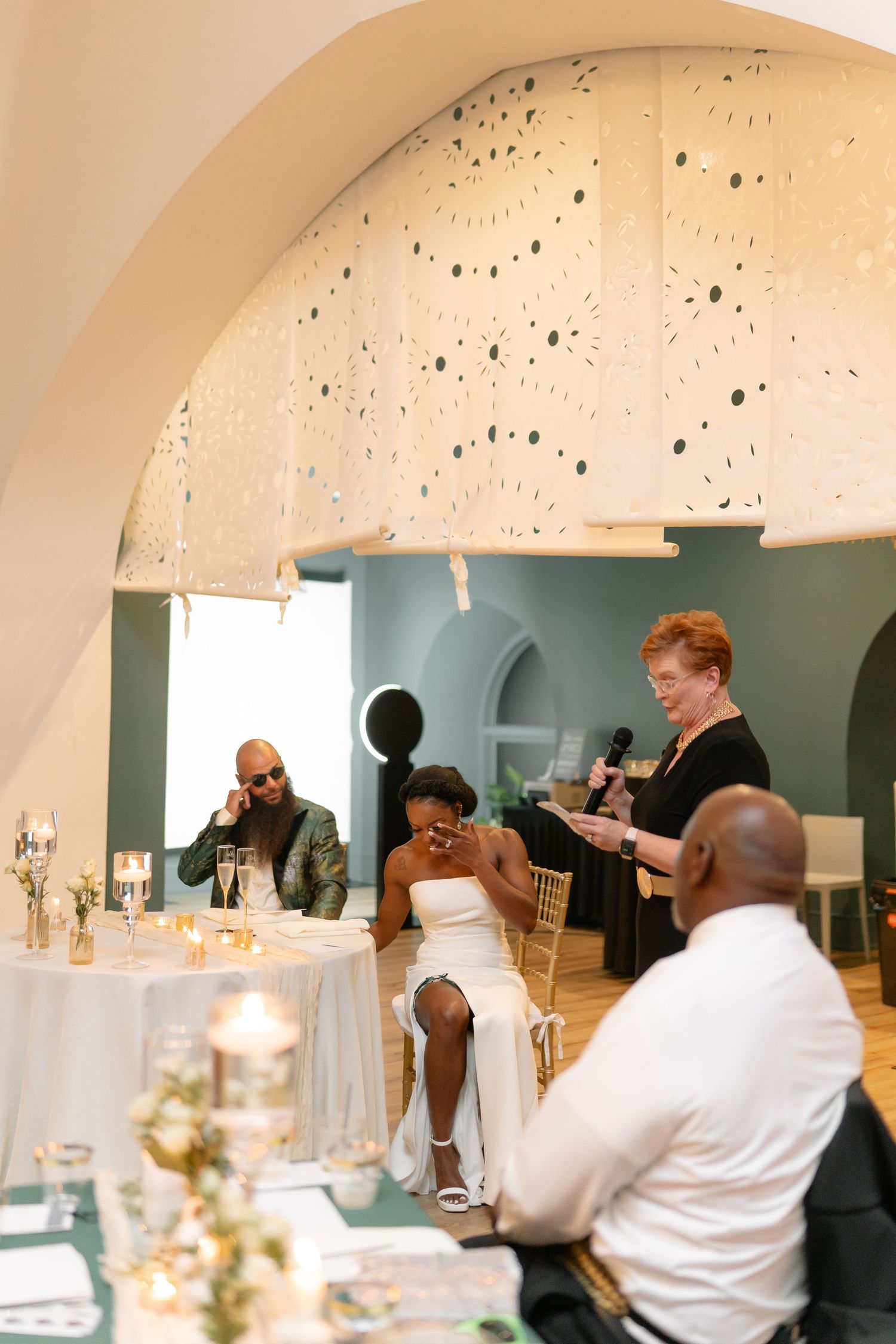 Wedding reception guests celebrate at decorated tables under an arched ceiling with decorative lighting.