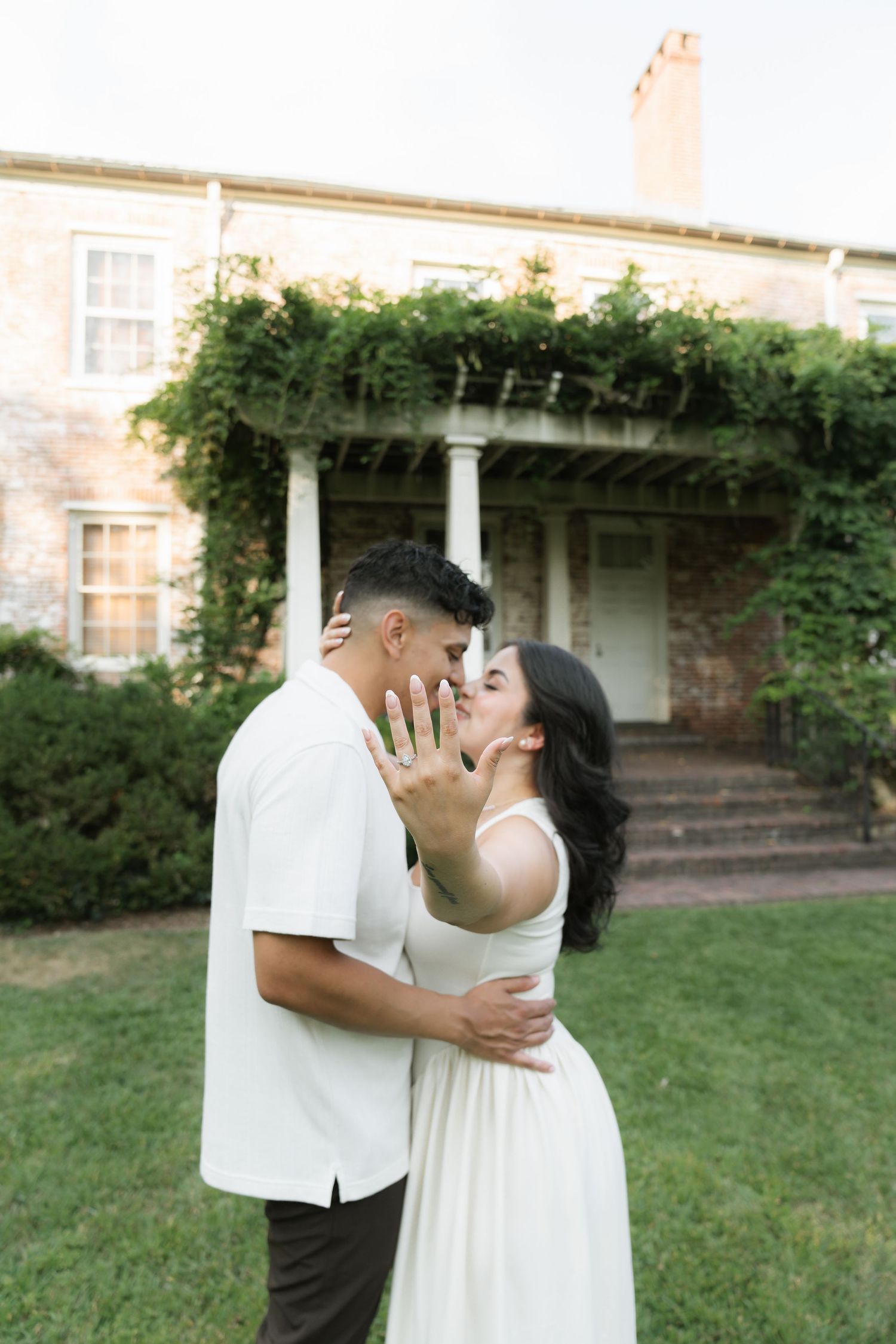 A couple in white attire share intimate moments in a garden setting with brick buildings in the background.