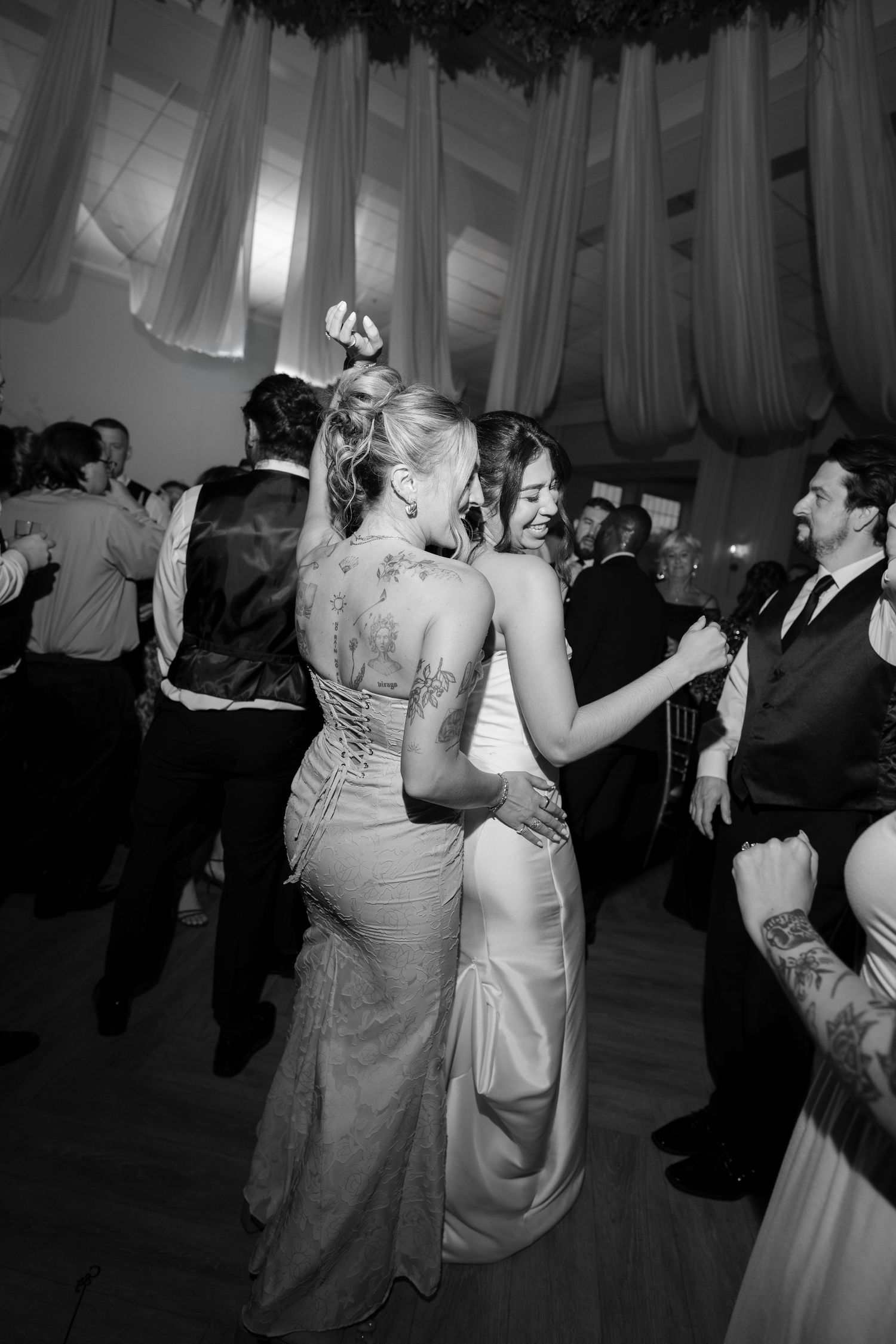 Black and white photo of bride and tattooed bridesmaid embracing on a crowded dance floor.