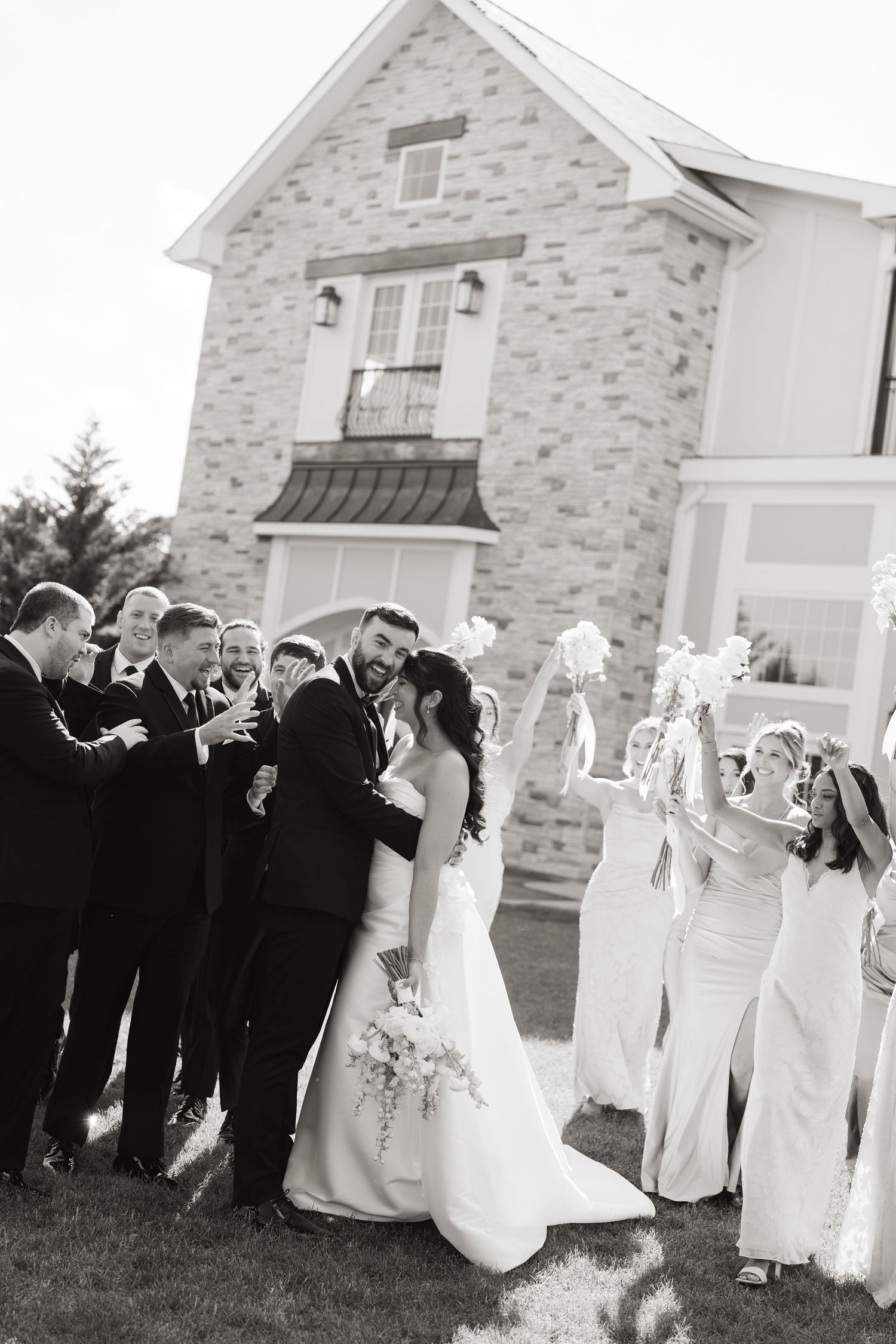 Black and white photo of wedding party celebrating with bride and groom kissing outside stone venue.