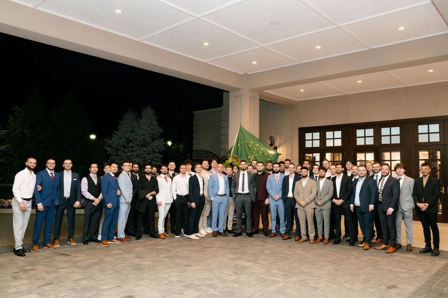 Large group photo of wedding guests lined up against a dark window wall in formal attire.