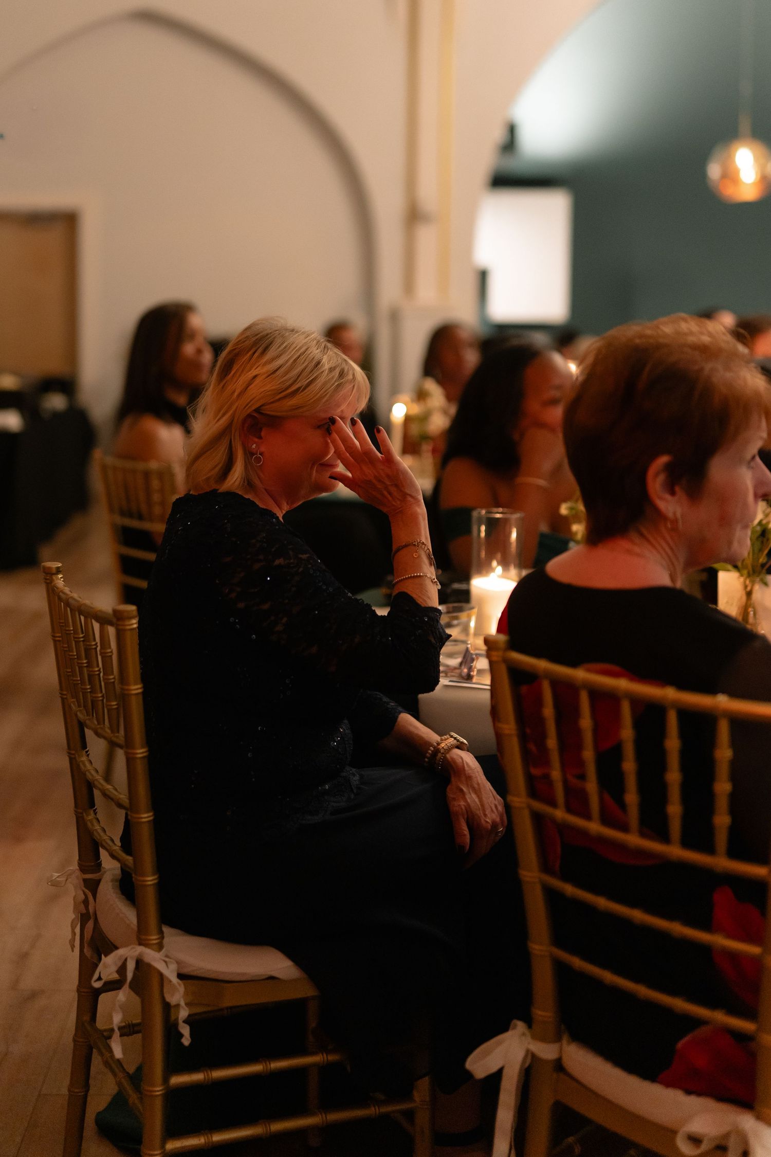 Wedding guests seated in gold chiavari chairs watch the festivities in a dimly lit reception space.