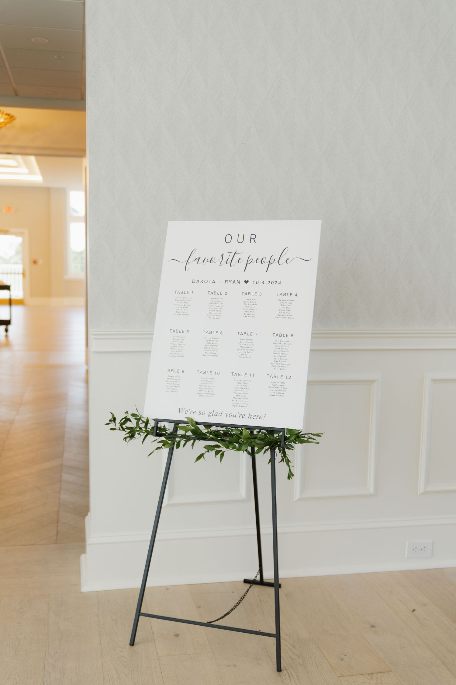 Wedding seating chart display on white easel with greenery decoration against light colored wall in elegant venue.