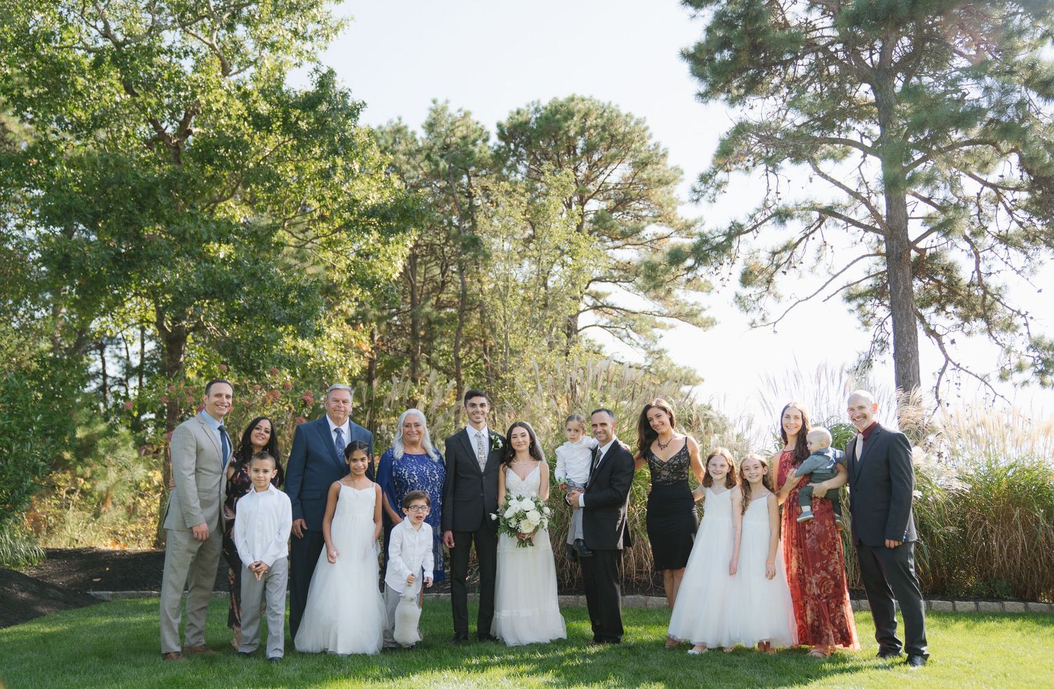 Wedding party members gather outdoors for group photos in a scenic garden setting with tall trees in the background.
