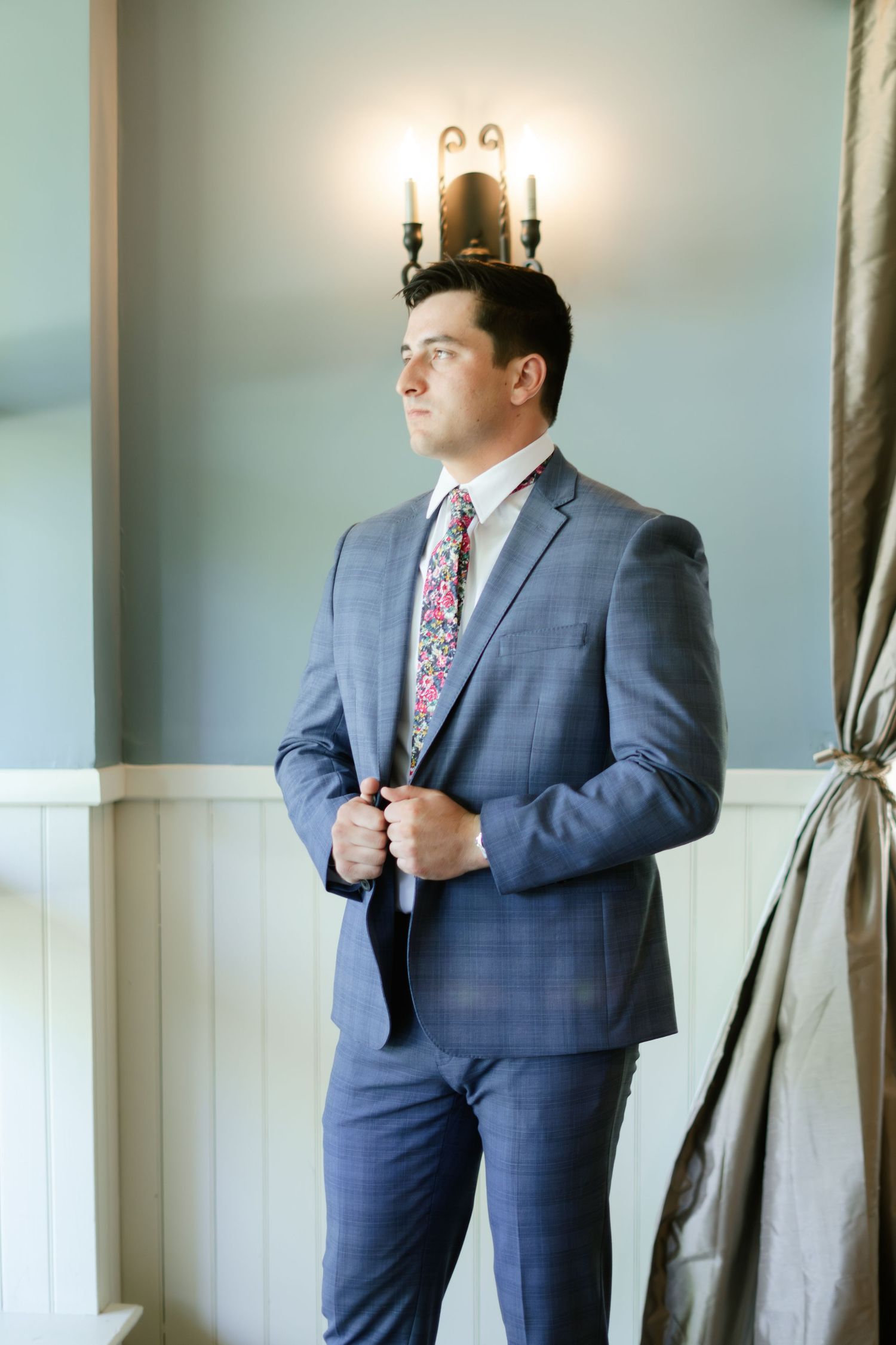 A person in a blue suit adjusts their tie while standing in an elegant room with curtains.