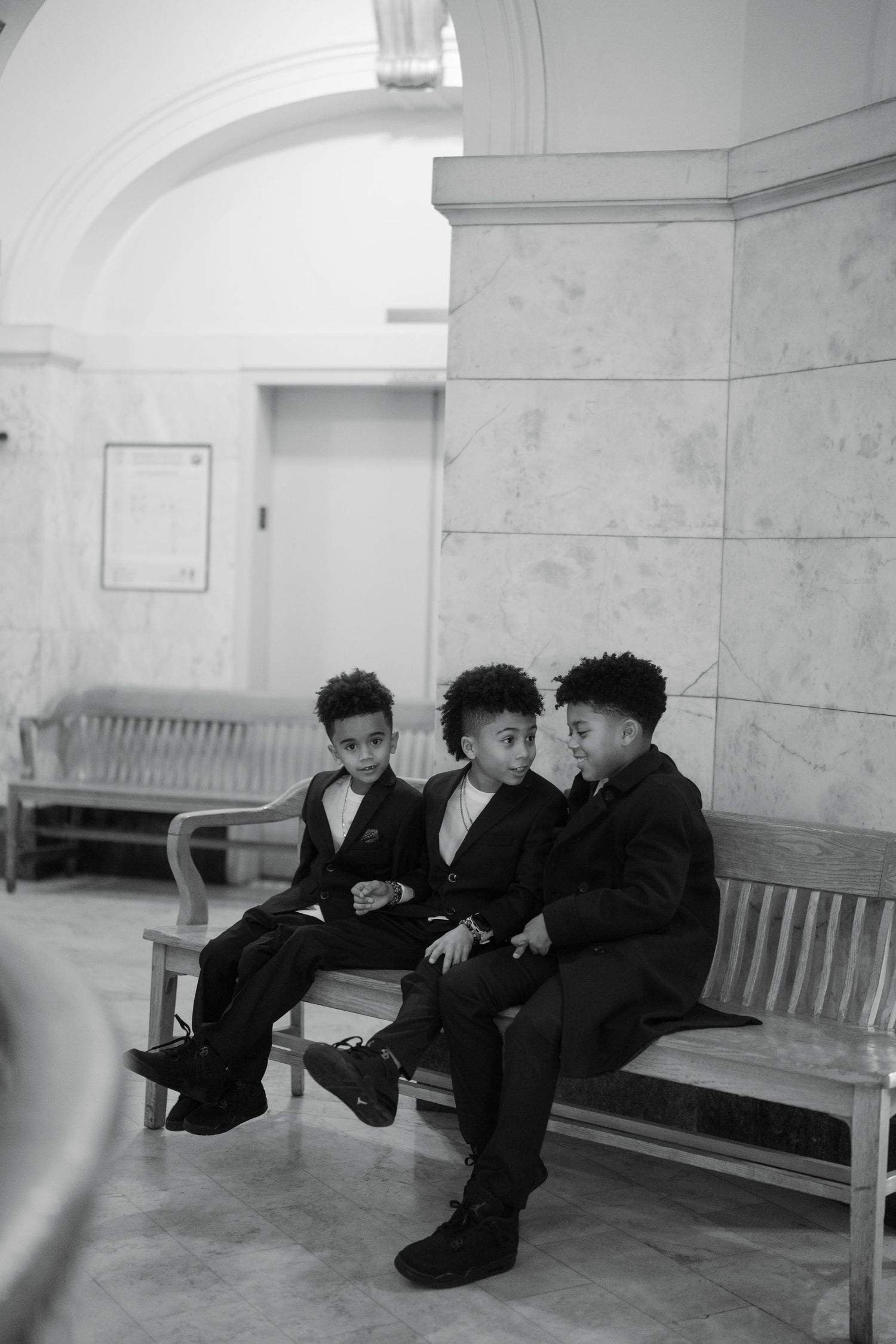 Three young boys in formal black suits sitting on a wooden bench in a marble hallway.