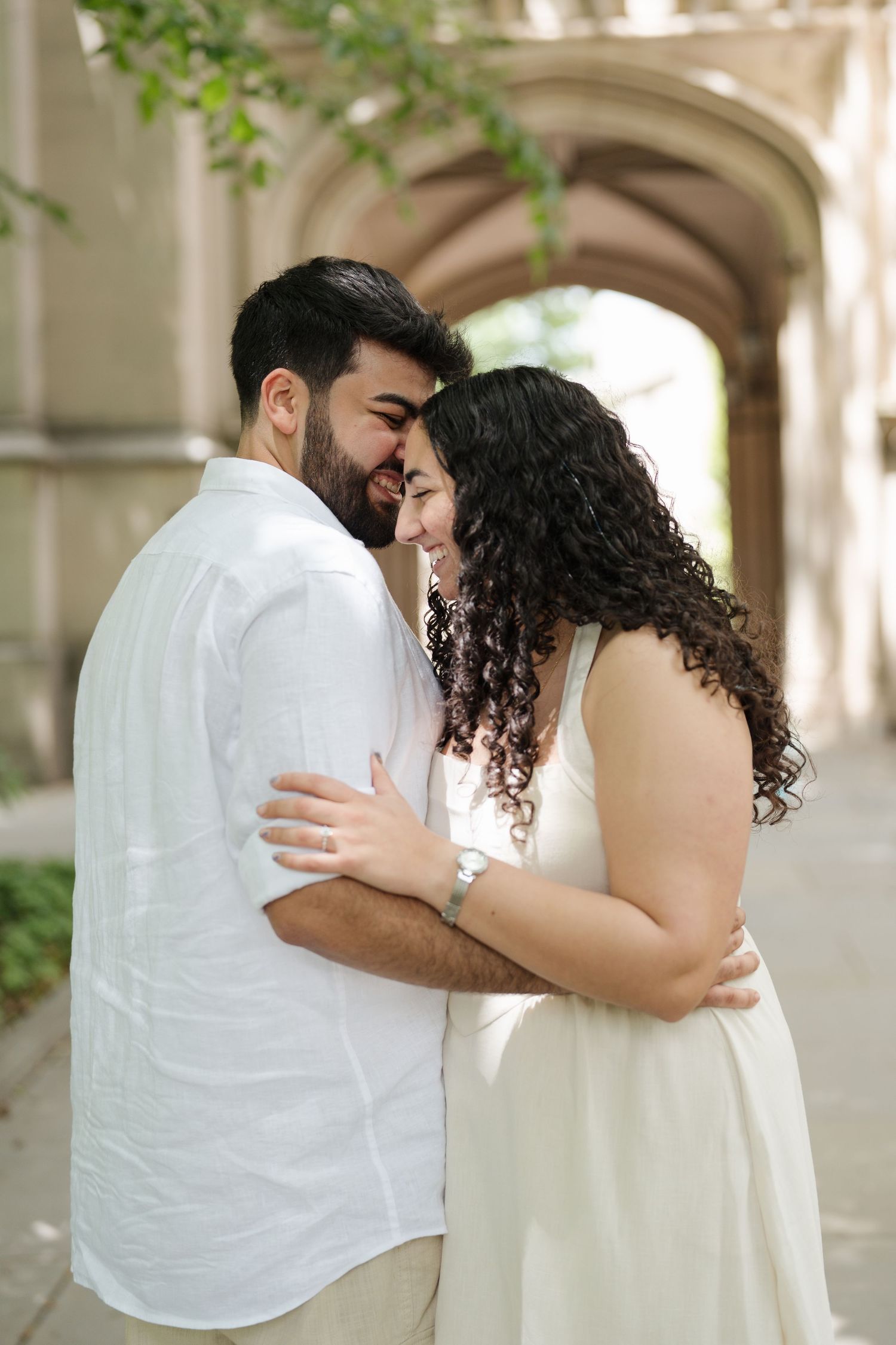A couple in white outfits embraces and smiles at each other in front of a stone archway outdoors.