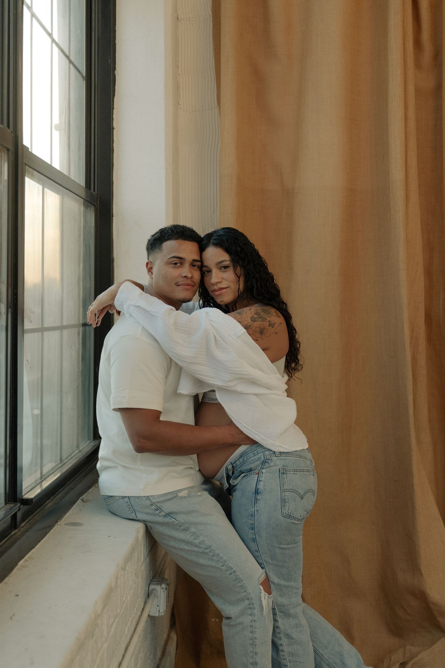 A couple in matching white tees and jeans pose together by a sunlit window with beige curtains.