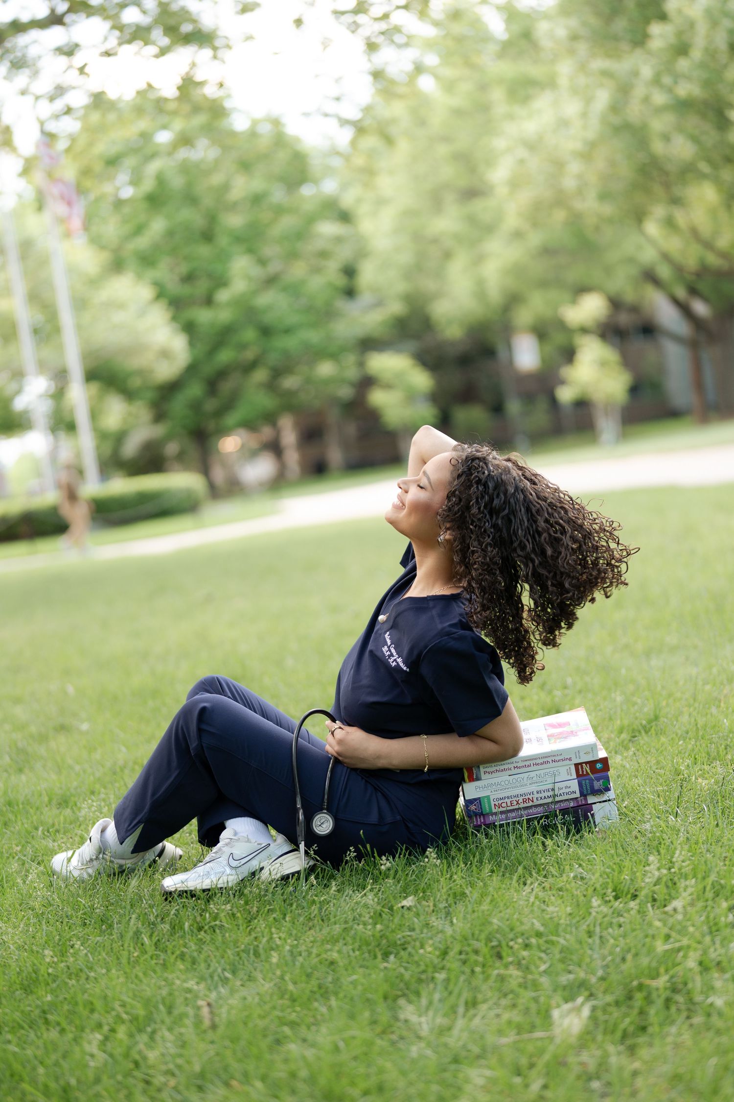 Nursing student in scrubs sits on grass with stethoscope and textbooks outdoors.