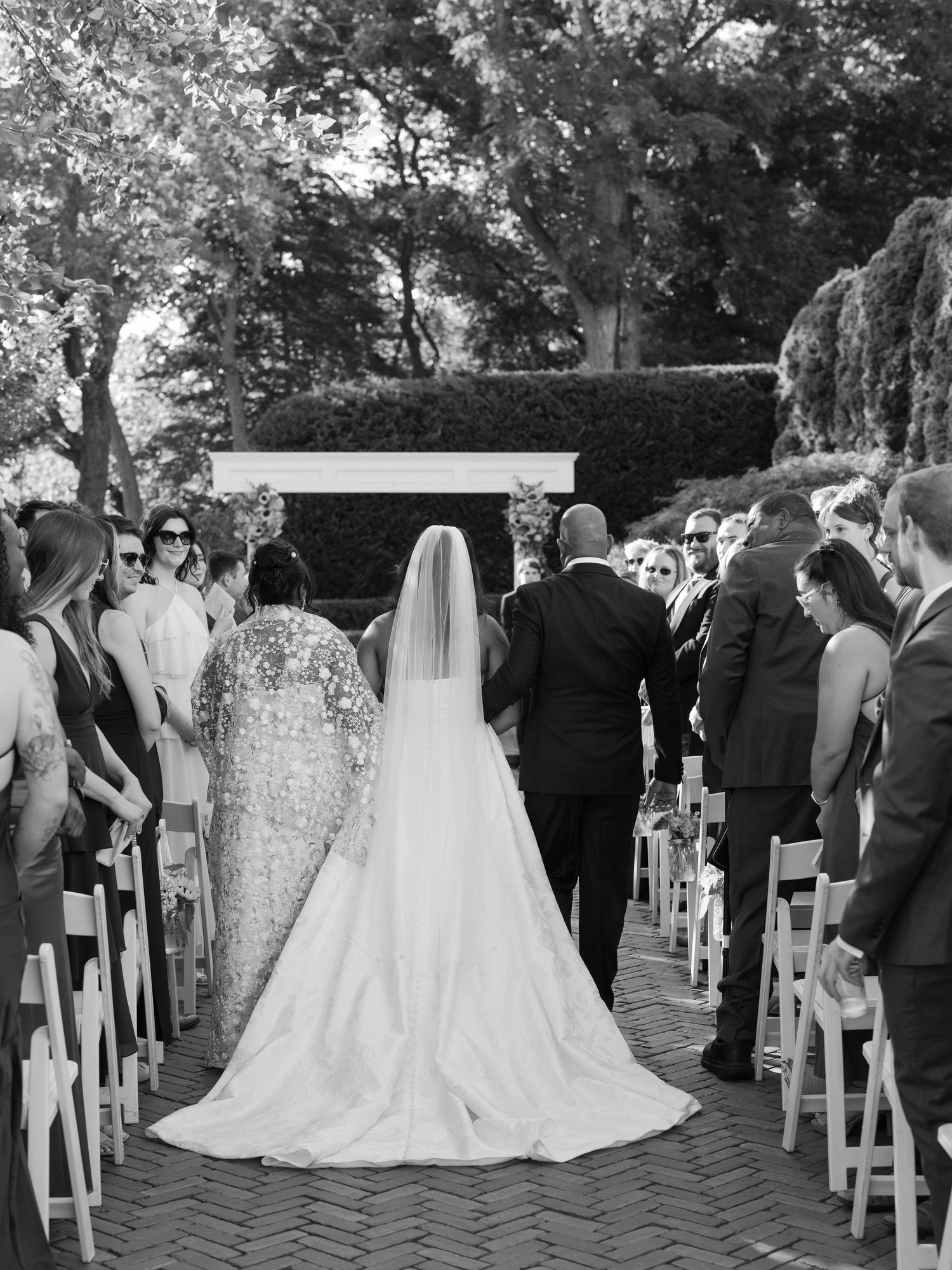 Black and white photo of a bride walking down the aisle at an outdoor wedding ceremony with seated guests.