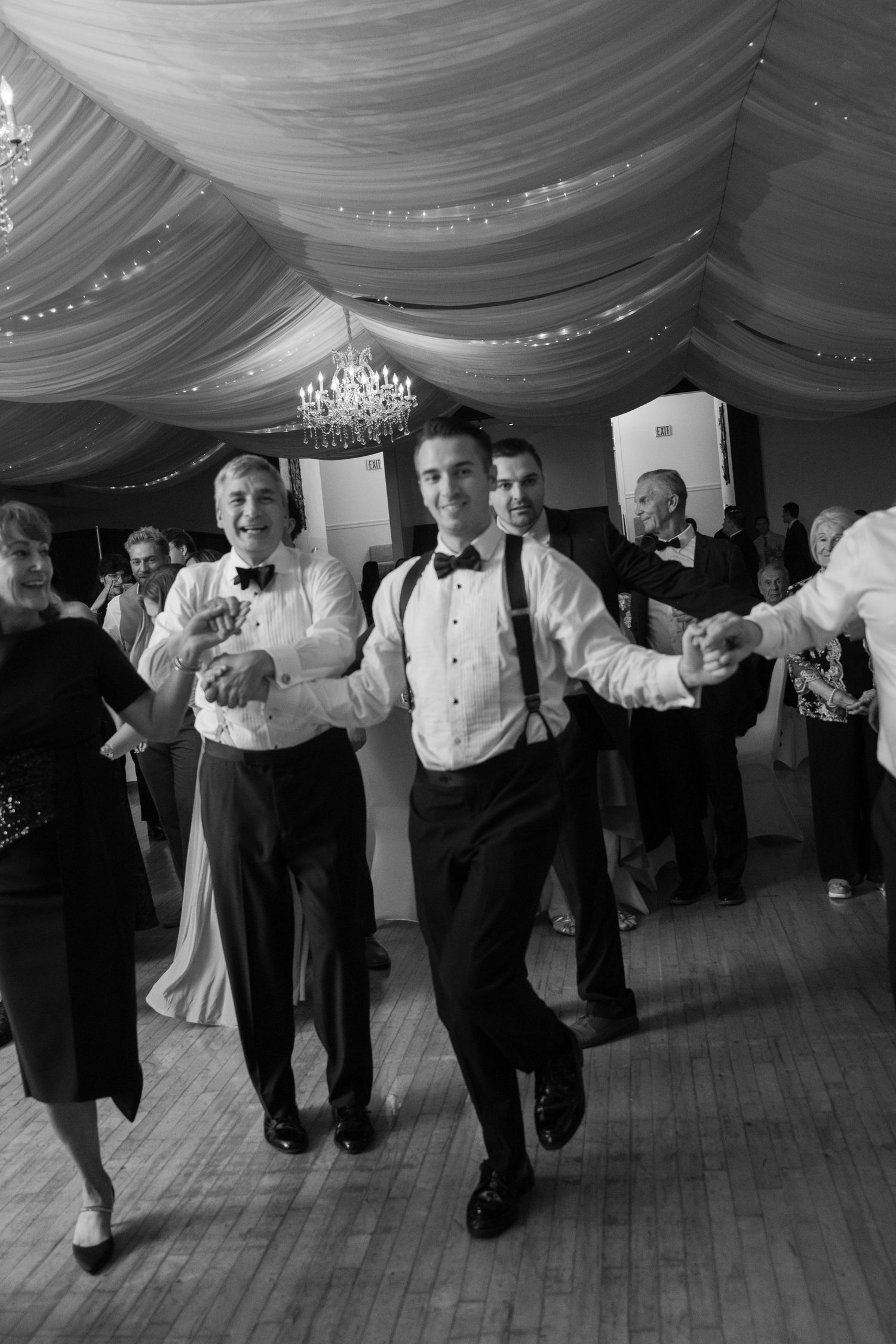 Black and white photo of groomsmen dancing energetically at elegant wedding reception venue.