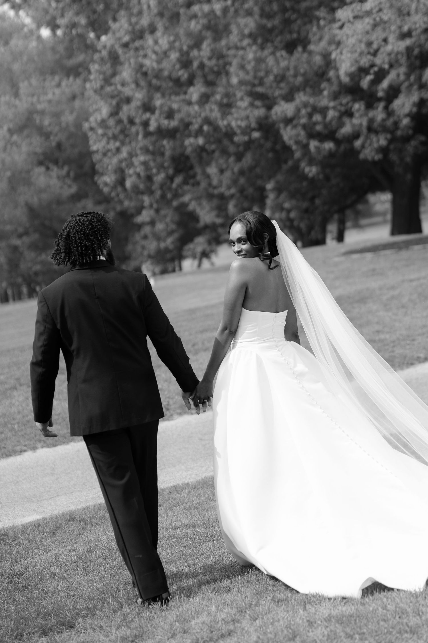 Black and white photo of a bride and groom holding hands while walking through a lush green park on their wedding day.