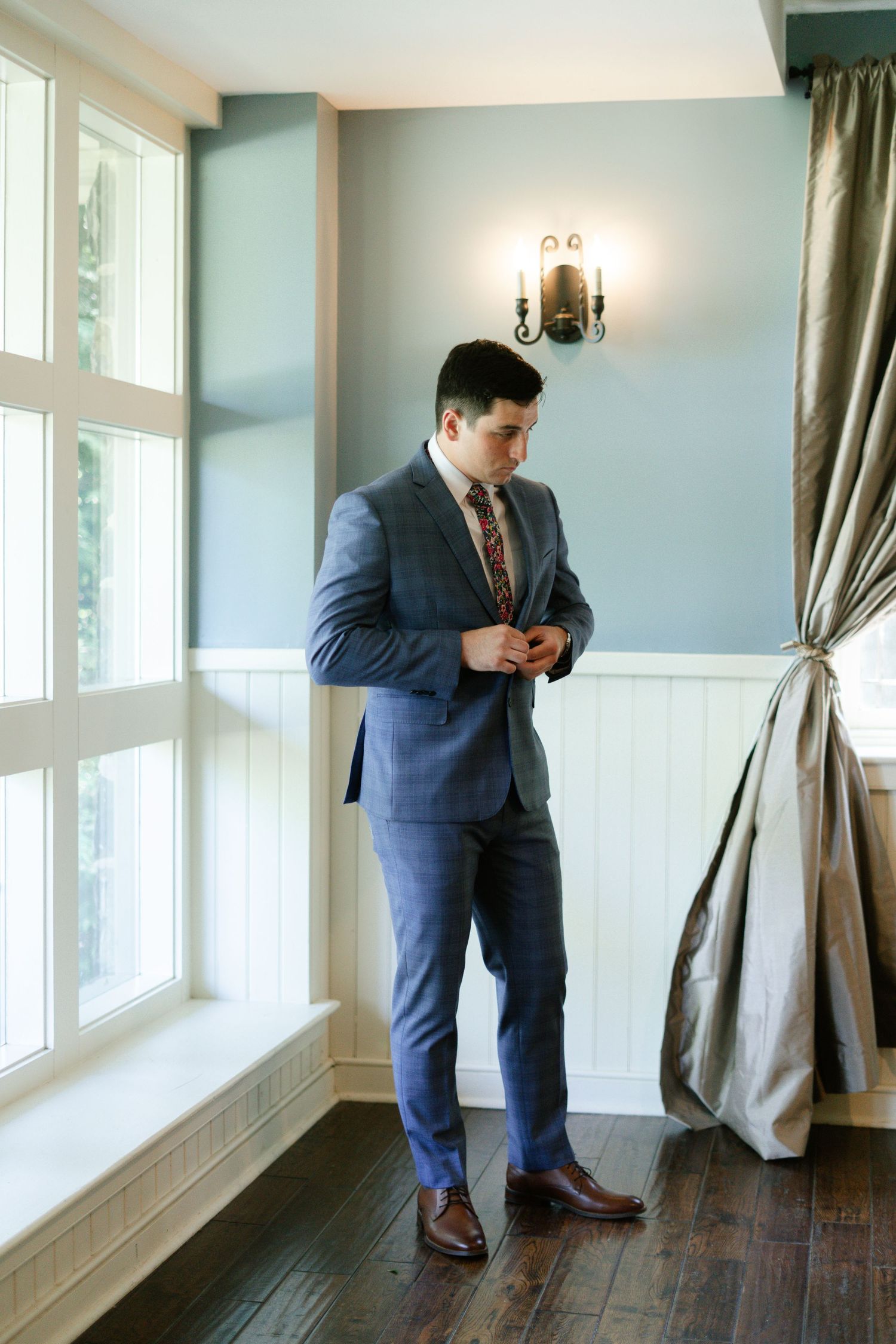A person in a blue suit and tie adjusts their jacket while standing by large windows in an elegant room with sage walls.