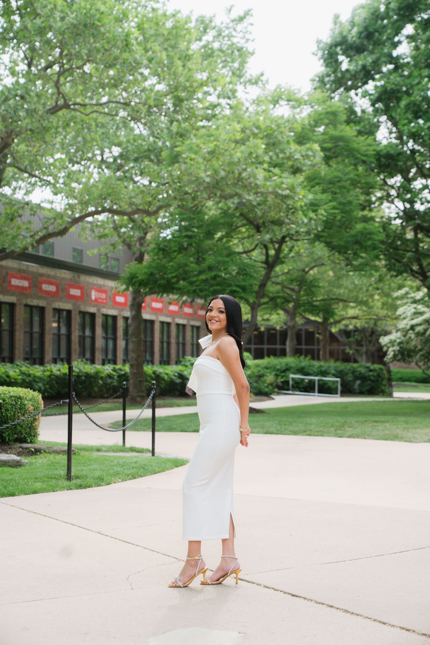 Graduate walking across a sunny campus sidewalk in white dress.