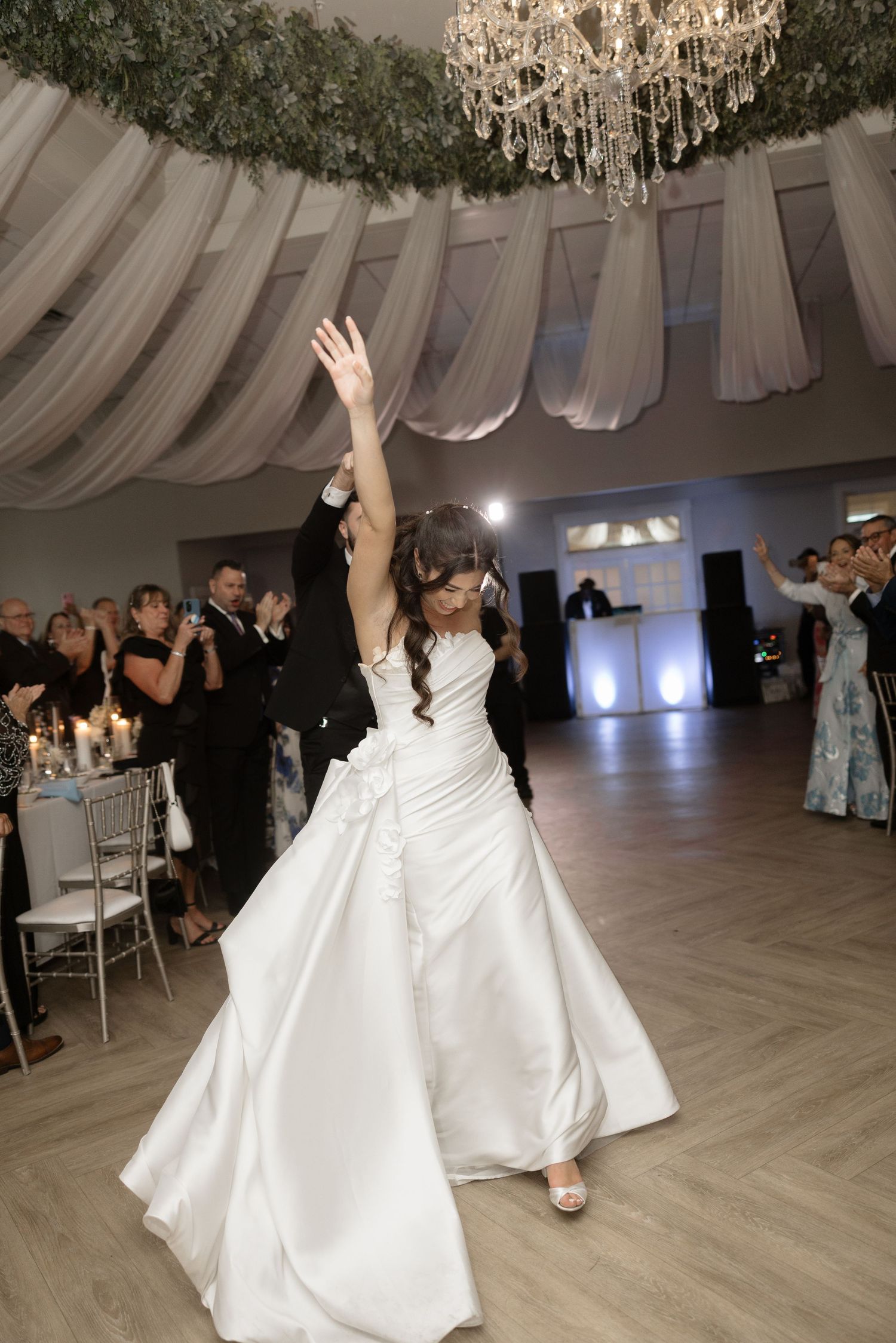 Bride in a white satin gown raises her arm joyfully during first dance at wedding reception.