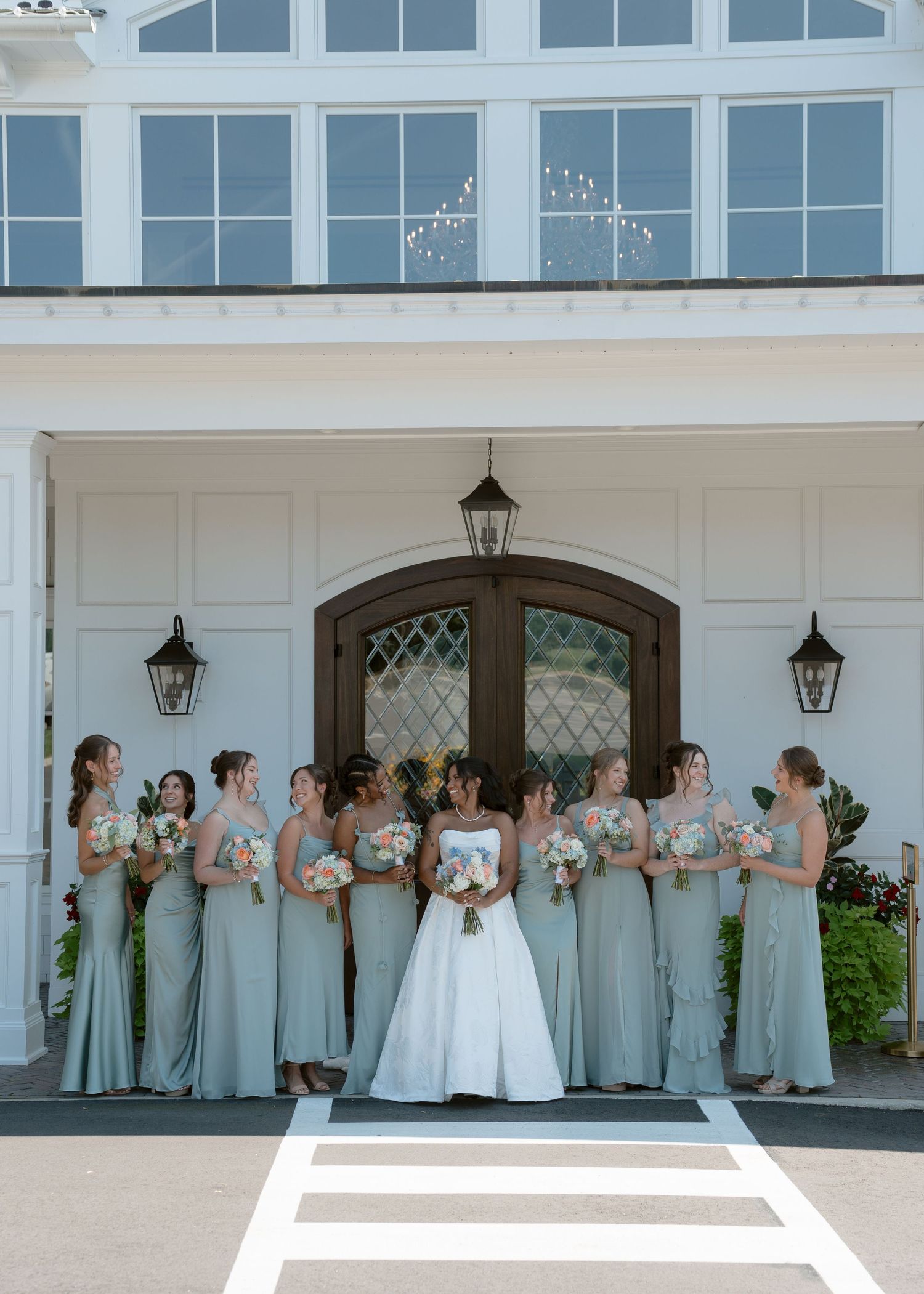 Bride in white gown with bridesmaids in sage green dresses holding bouquets outside white venue with arched doors.