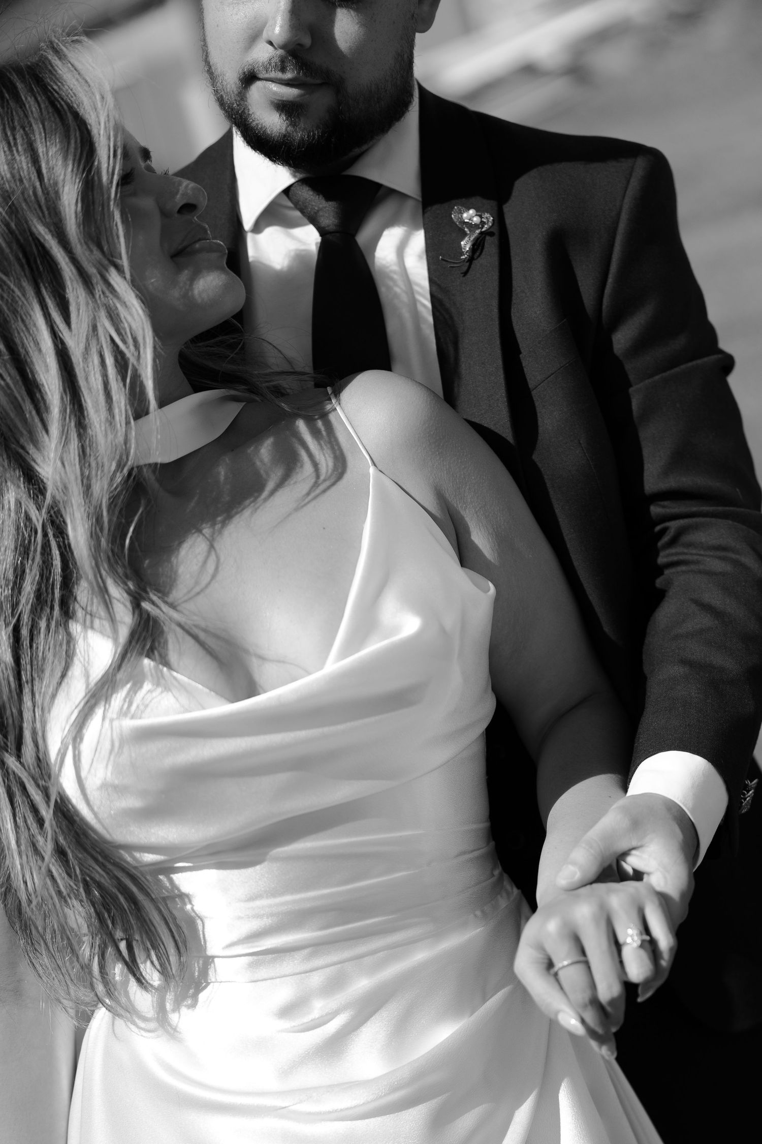 Black and white portrait of bride in satin gown holding grooms hand, wedding ring visible, romantic outdoor setting.