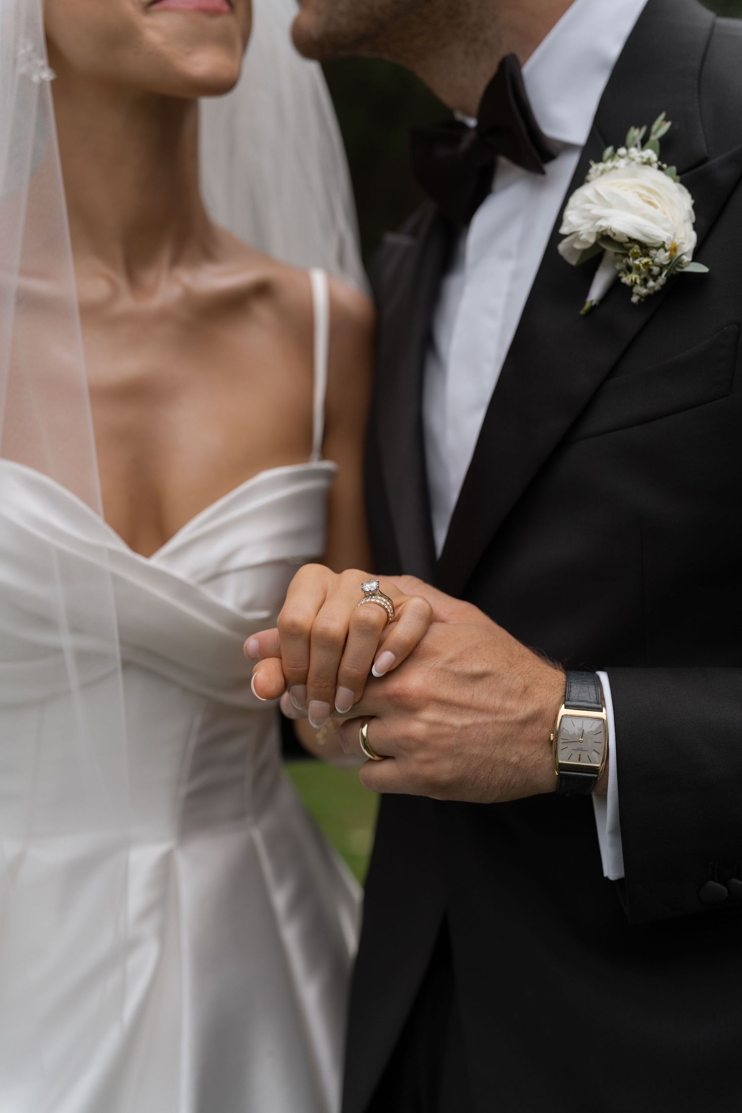 Close-up of bride and groom holding hands showing diamond engagement ring and groom's boutonniere.