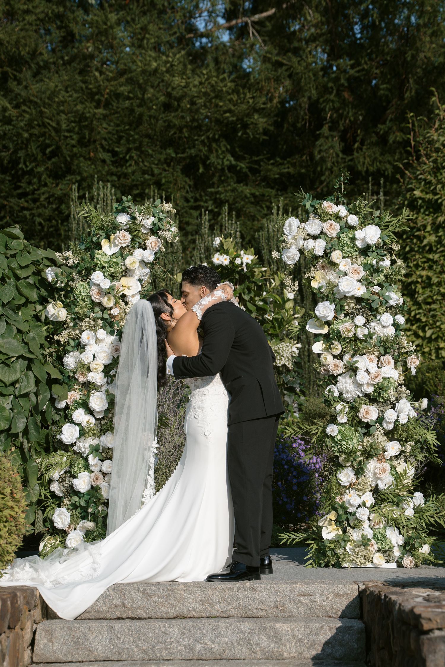 Bride and groom share first kiss under a stunning double floral arch with white and blush roses outdoors.