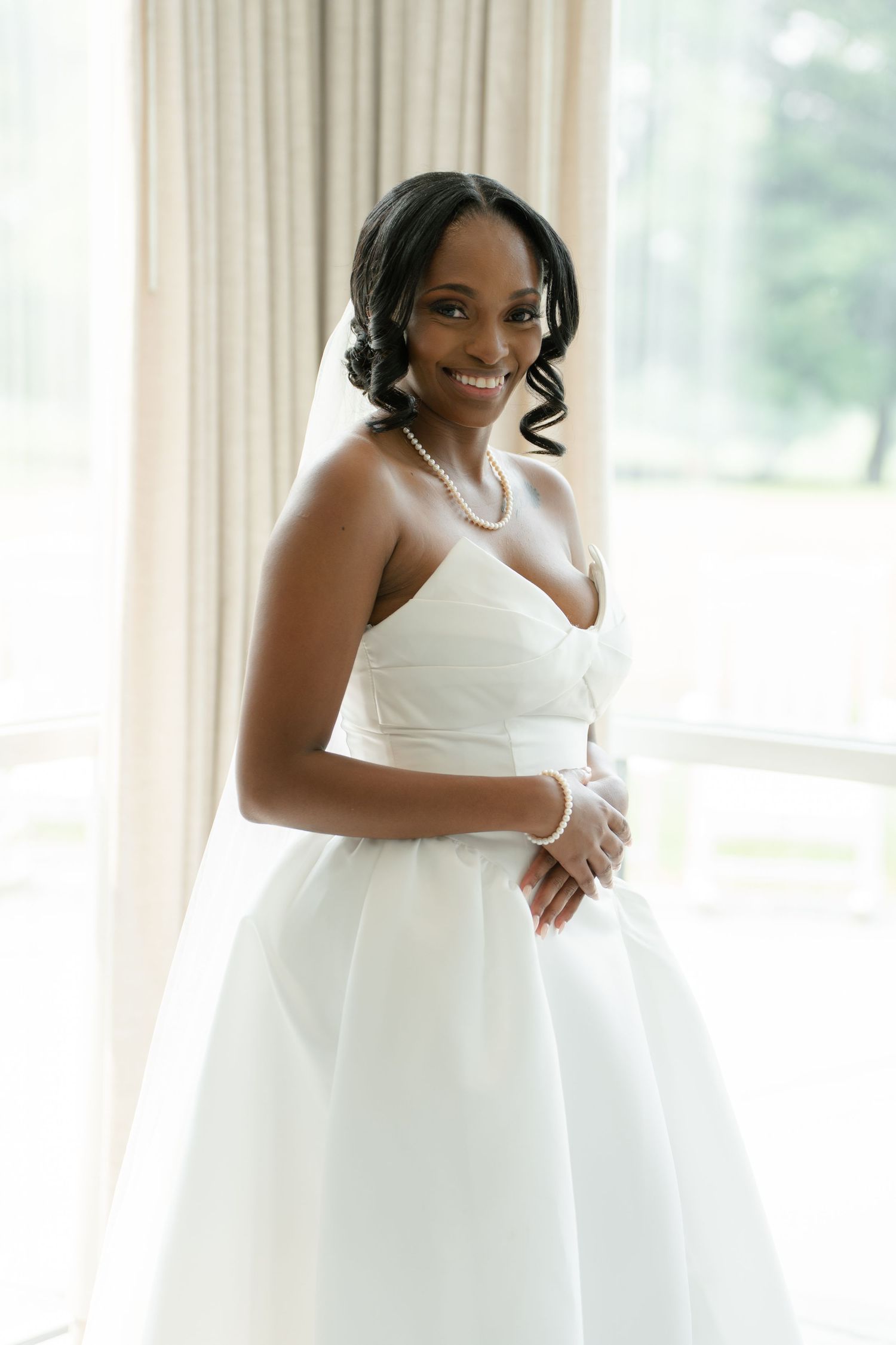 Smiling Black bride in strapless white ball gown with pearl necklace and veil poses by bright window.