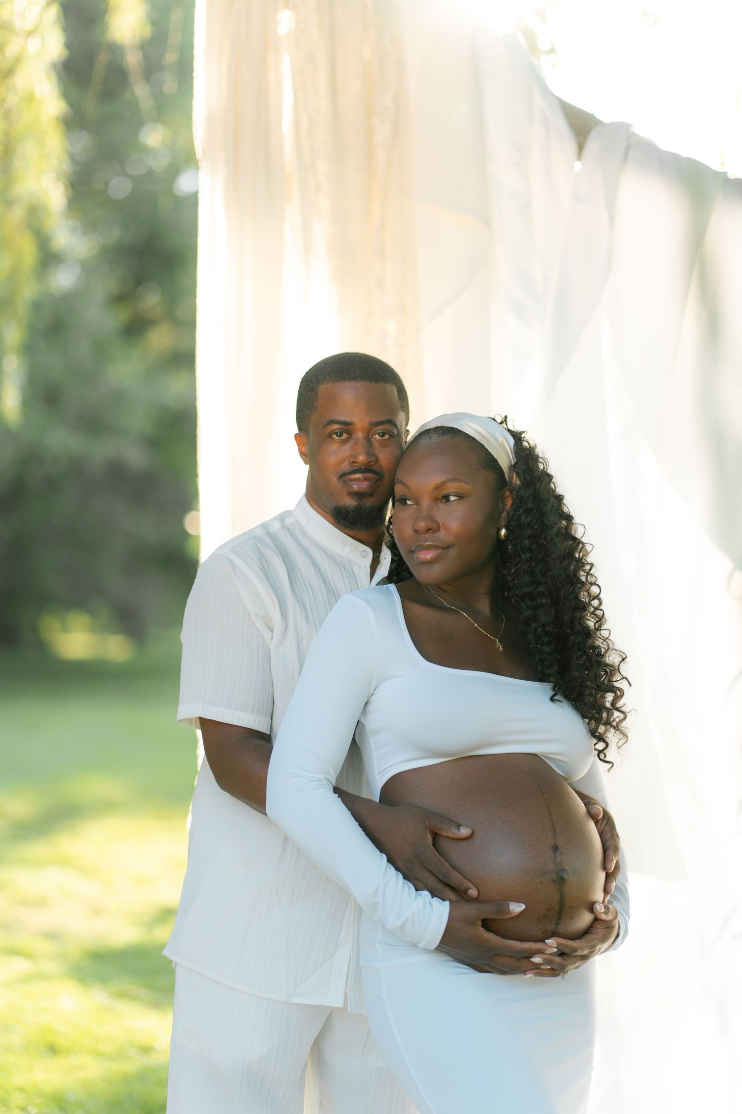 A pregnant couple in white outfits pose outdoors, the man embracing the woman from behind as she cradles her baby bump.