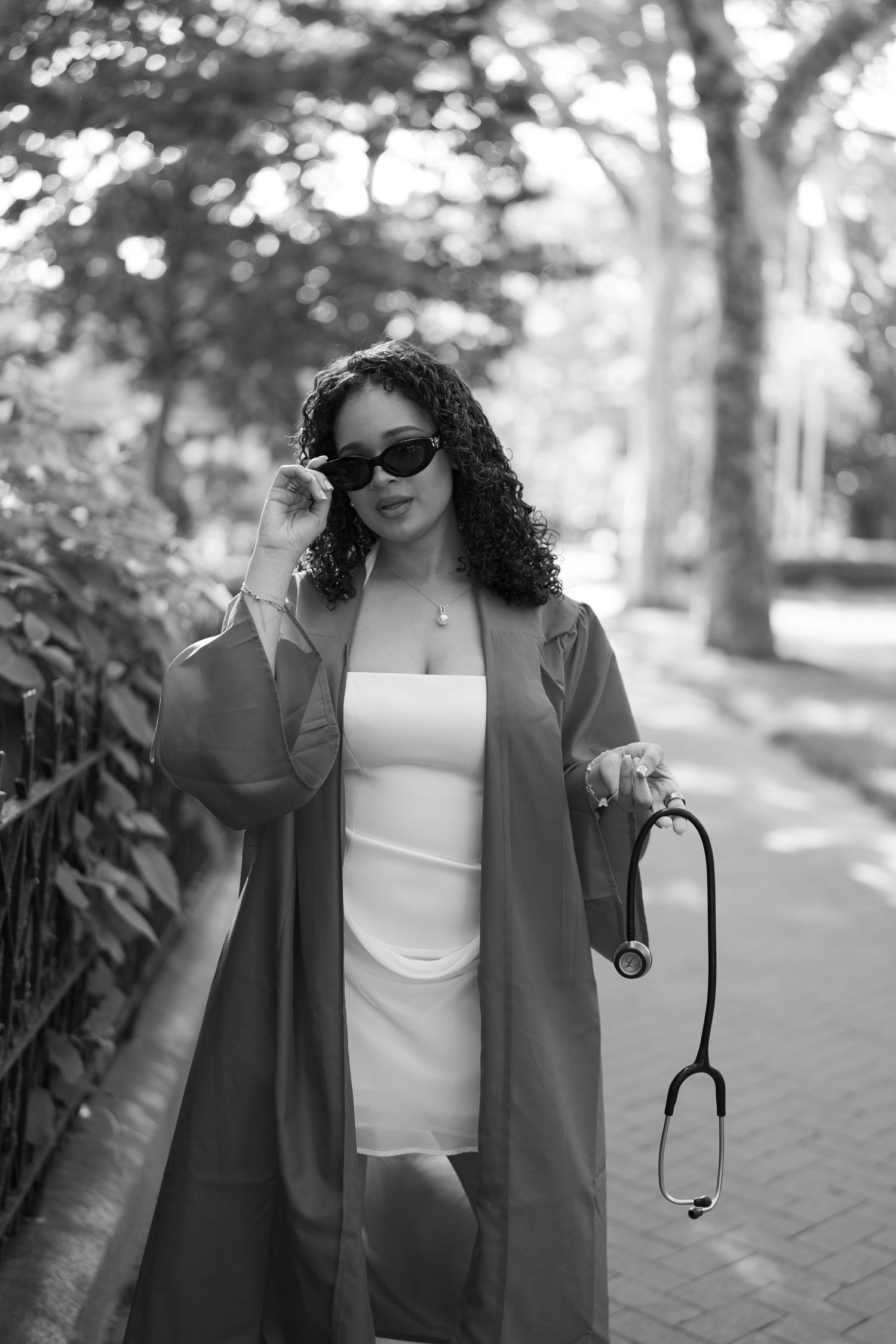 Black and white photo of young woman in graduation gown holding stethoscope and sunglasses outdoors.