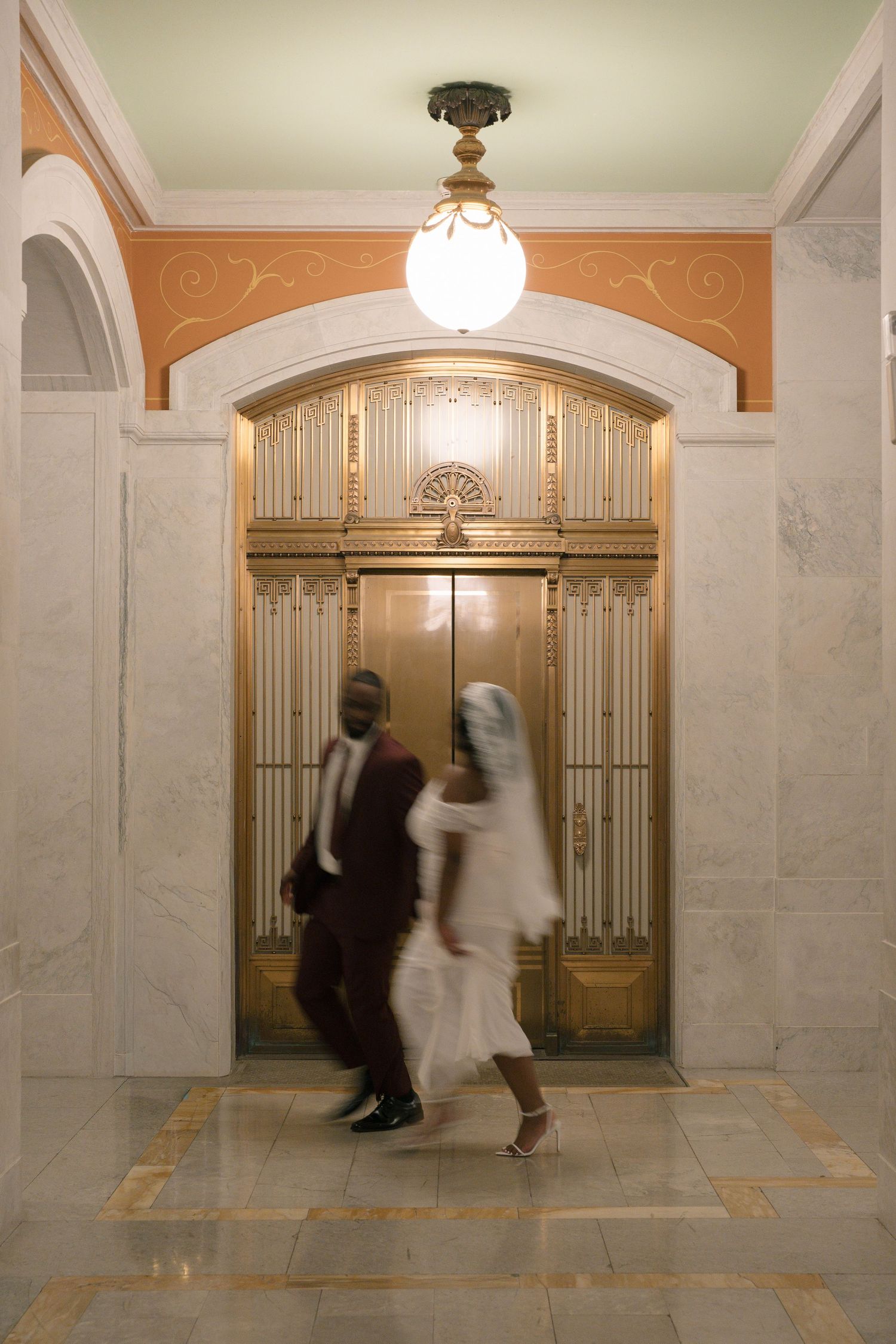 Blurred motion of a couple in formal wear walking past an ornate gold elevator in a marble lobby.