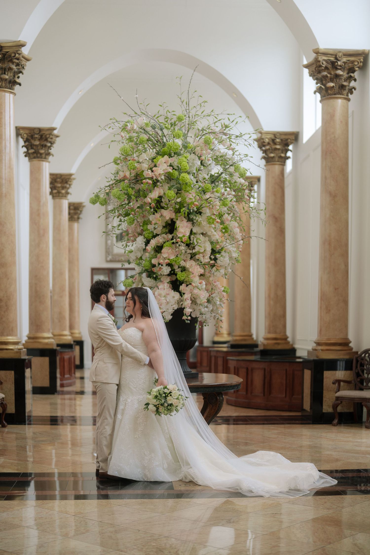 Romantic wedding couple embraces in an elegant marble columned hall with ornate floral arrangements and arched architecture.