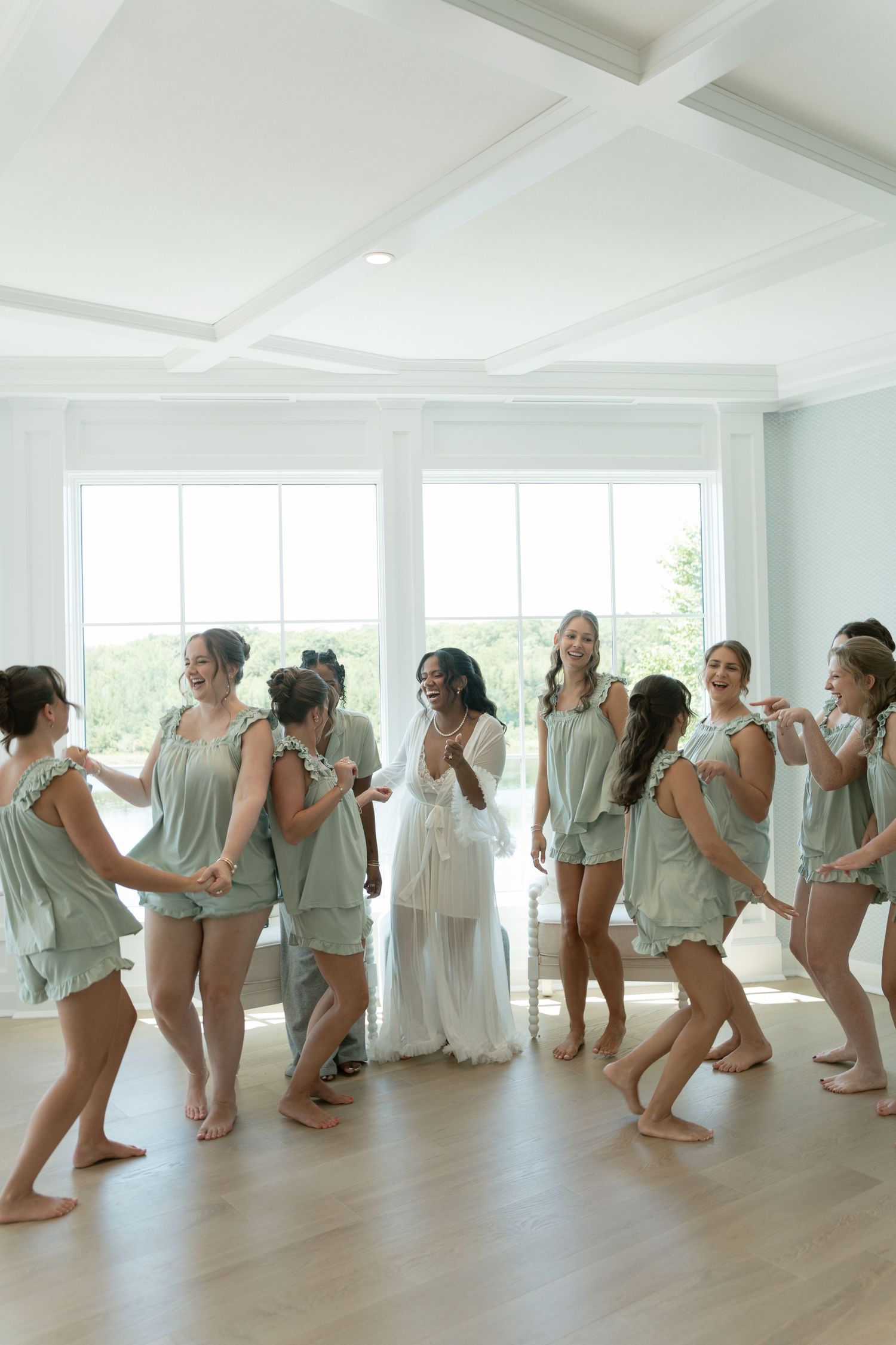 Bride in white robe surrounded by bridesmaids in sage green matching sets dancing together in a bright room.