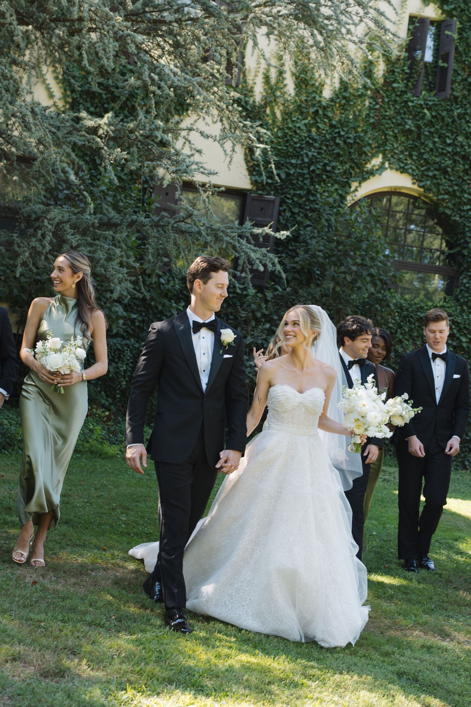 Wedding party walking and laughing together outdoors in formal attire with white floral bouquets.