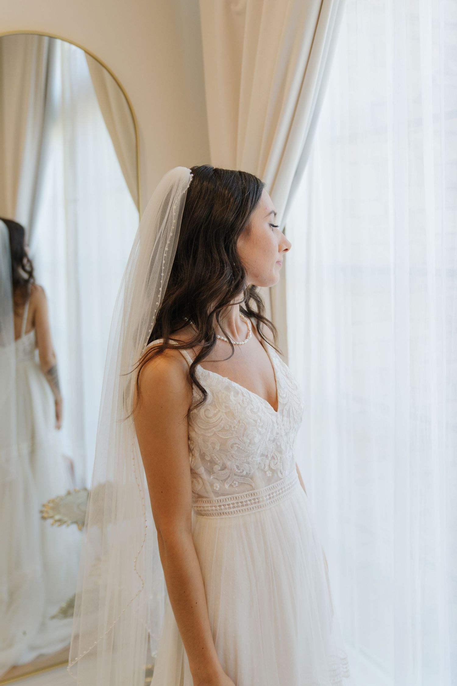 A bride in a flowing white lace wedding dress and veil stands serenely by a sunlit window.