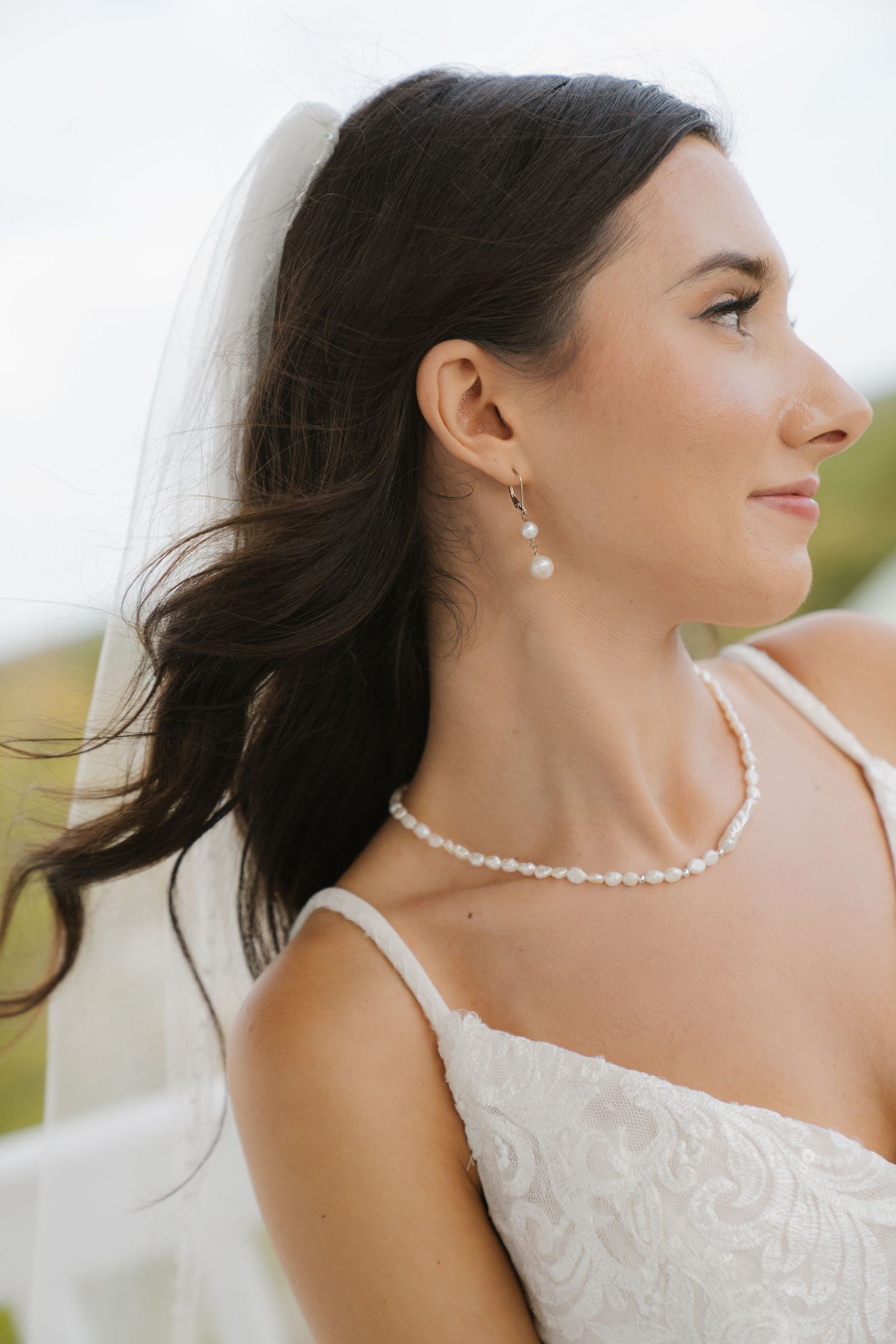 A bride in a white lace dress with pearl necklace and earrings smiles while looking to the side with windswept hair.