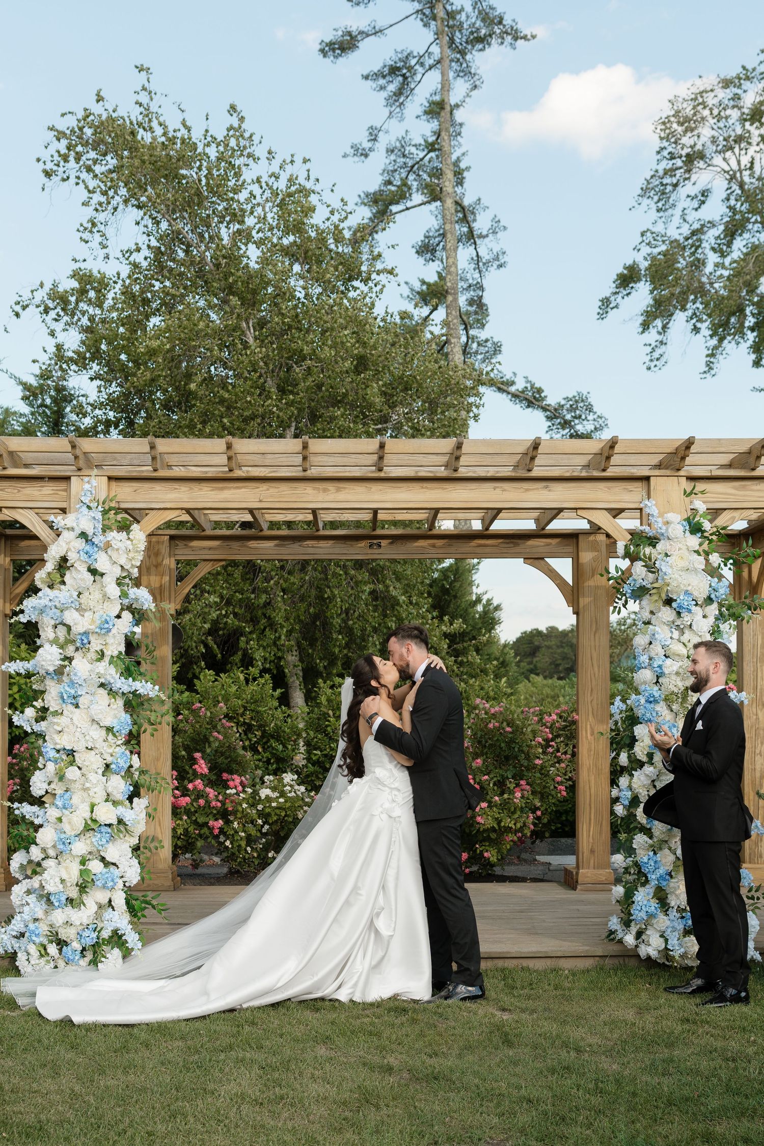 Bride and groom share first kiss under a wooden pergola adorned with blue and white flowers.