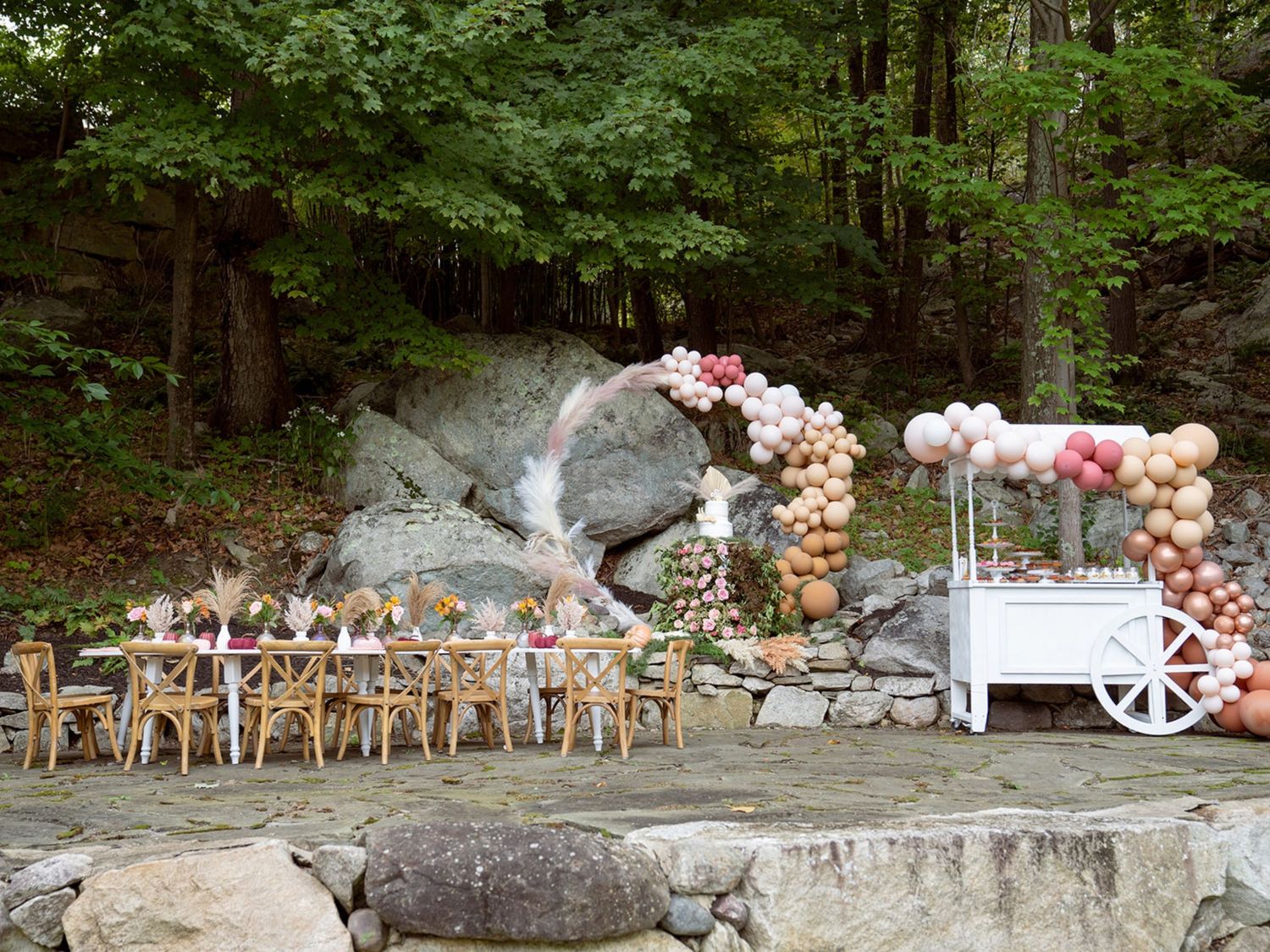 Outdoor wedding setup with balloon arch and wooden chairs against a rocky forest landscape.