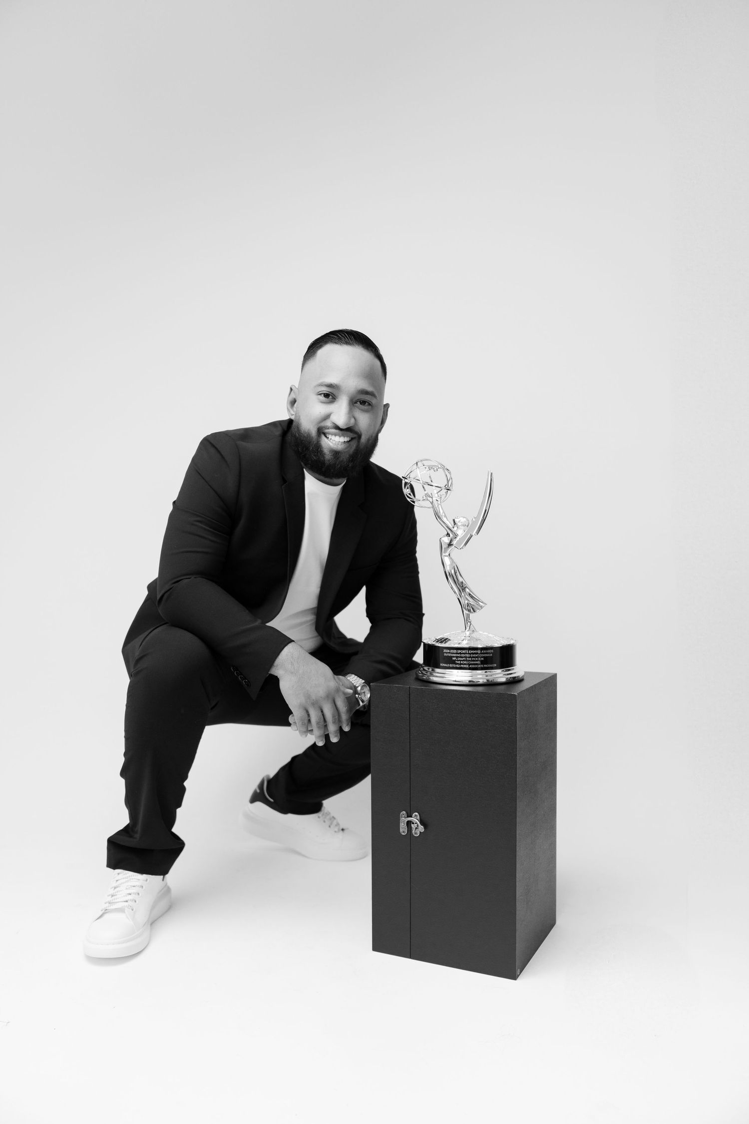 Professional in black suit posing with Emmy award trophy against white background.