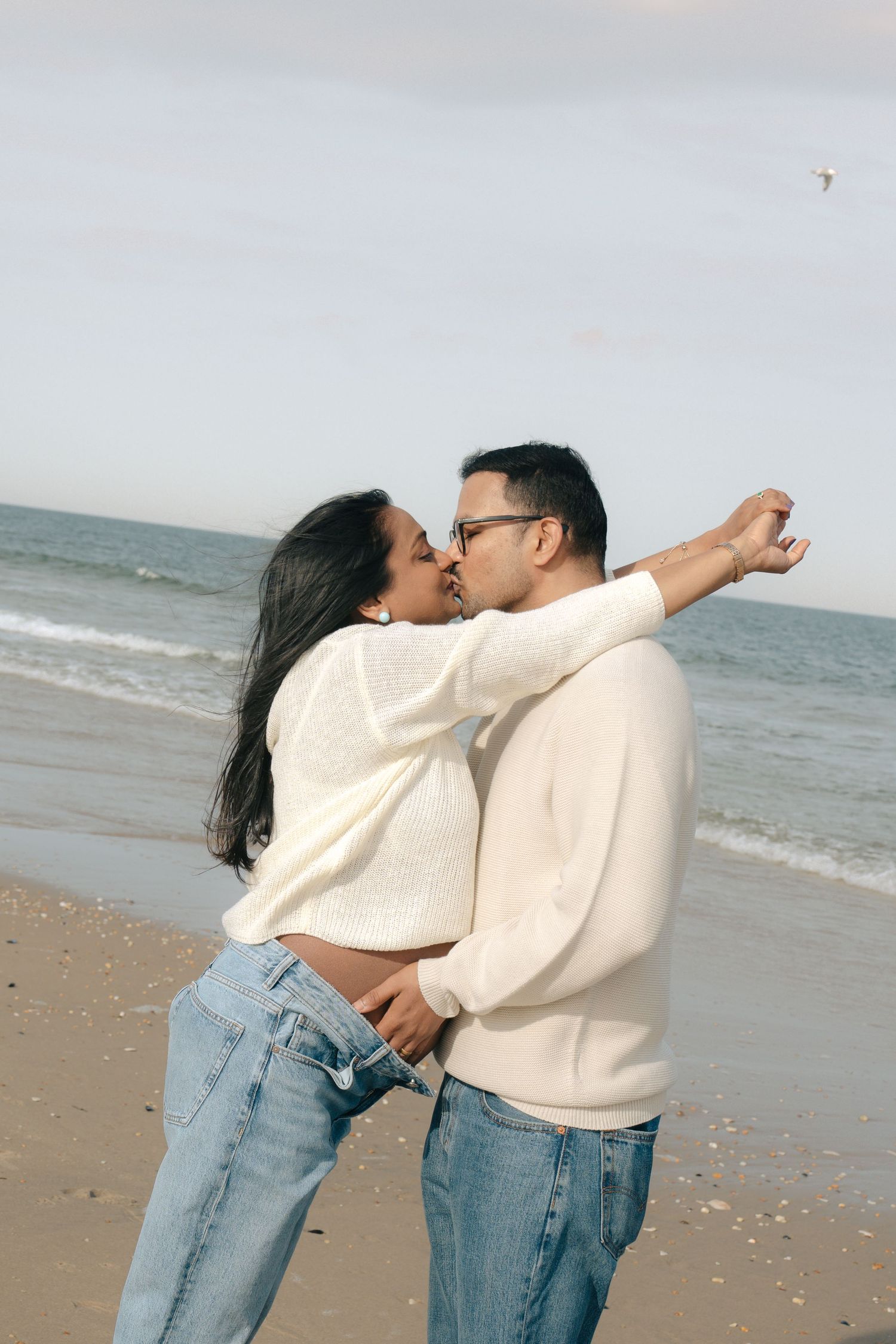 A couple wearing matching cream sweaters and jeans embrace playfully on a cloudy beach.