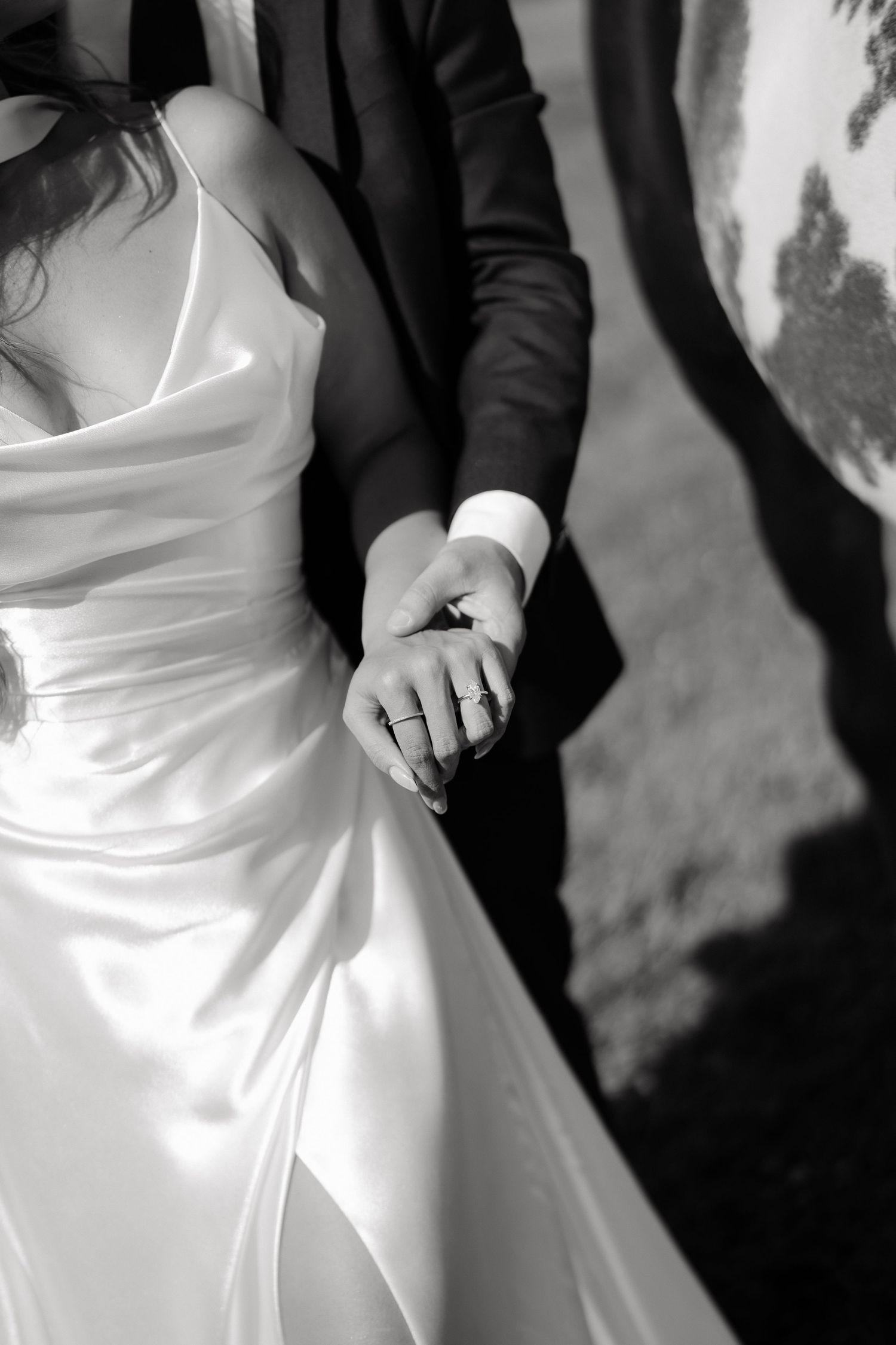 Black and white photo of bride and groom holding hands, showcasing wedding rings, bride in elegant satin gown.