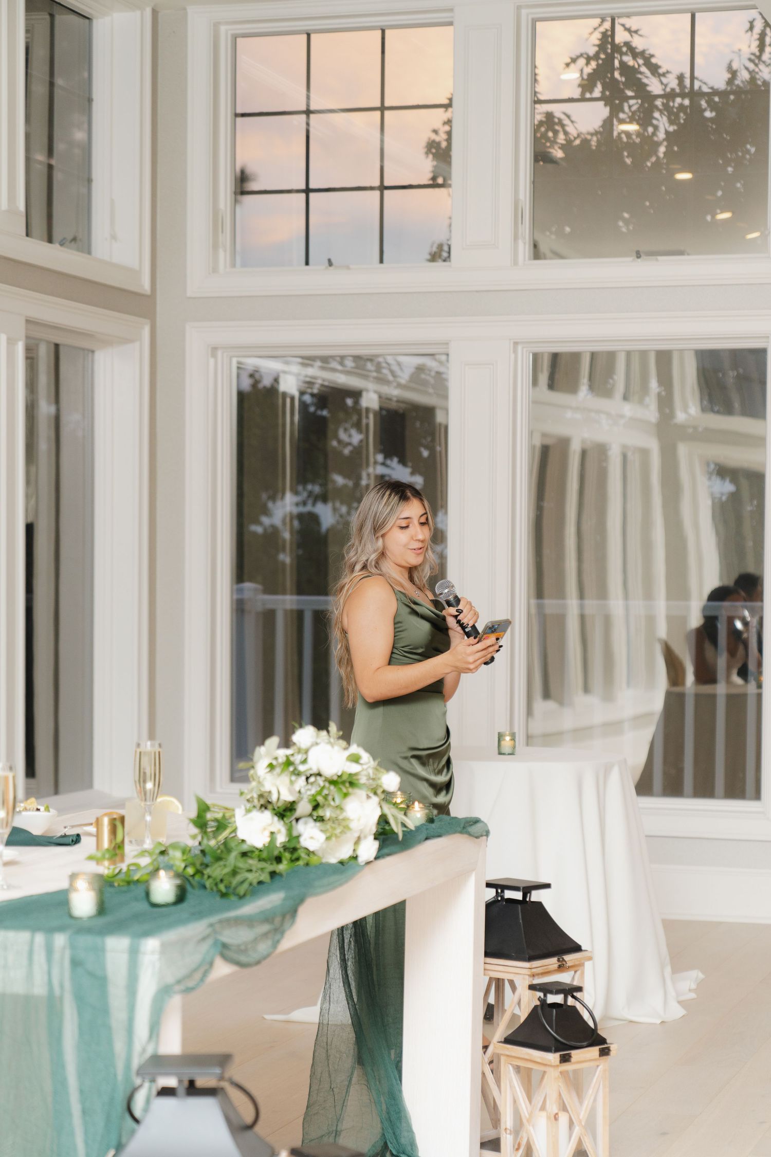 A green-dressed figure stands by an elegant table with white floral arrangements and teal decor in a sunlit room.